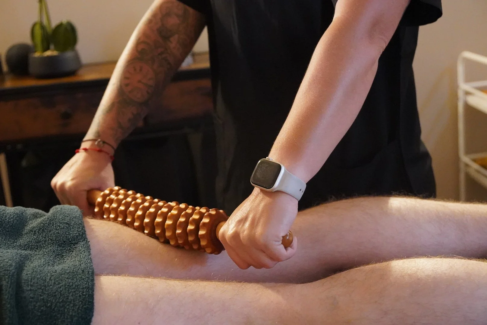A massage therapist uses a wooden massage roller on a client's leg. The therapist's arms are visible, with one having a tattoo sleeve and wearing a smartwatch. The background includes a potted plant and some furniture.