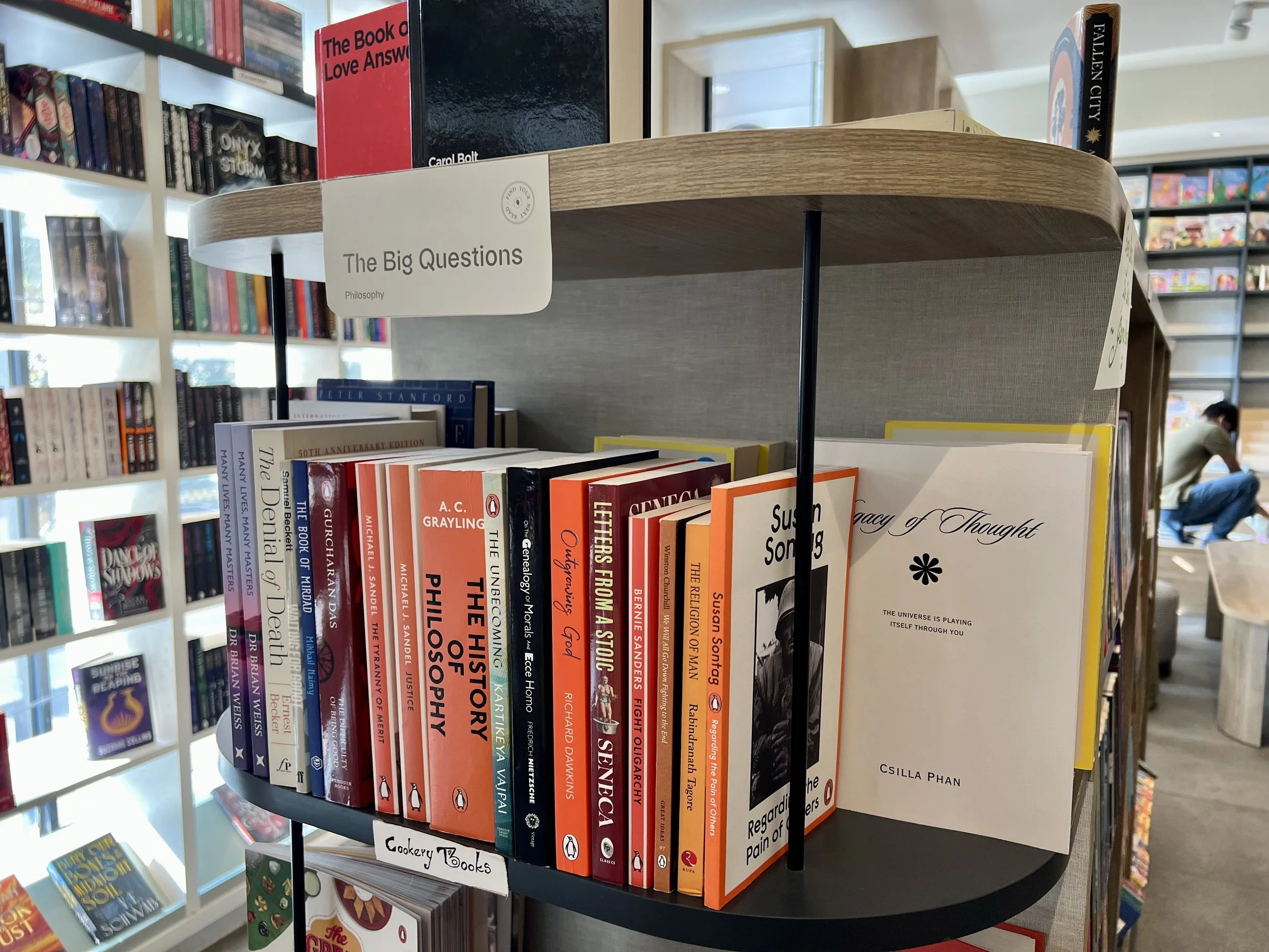 Bookshelf in a bookstore with titles on philosophy, history, and other genres, labeled 'Cookery Books' and 'The Big Questions' sign.