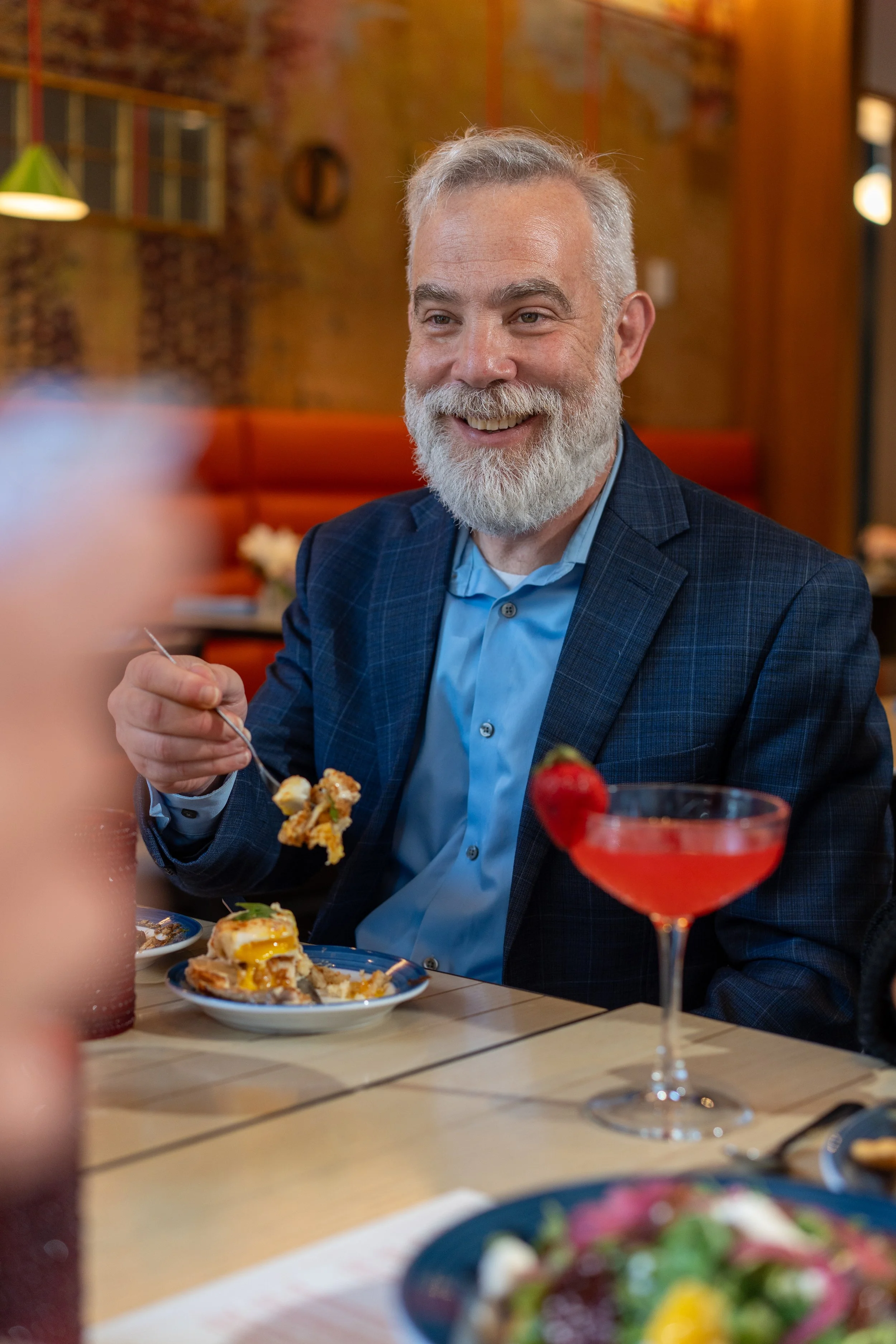 An older man with a white beard and gray hair wearing a blue checkered blazer and light blue shirt, smiling while eating at a restaurant. A plate with food and a red cocktail in a fancy glass are on the table.