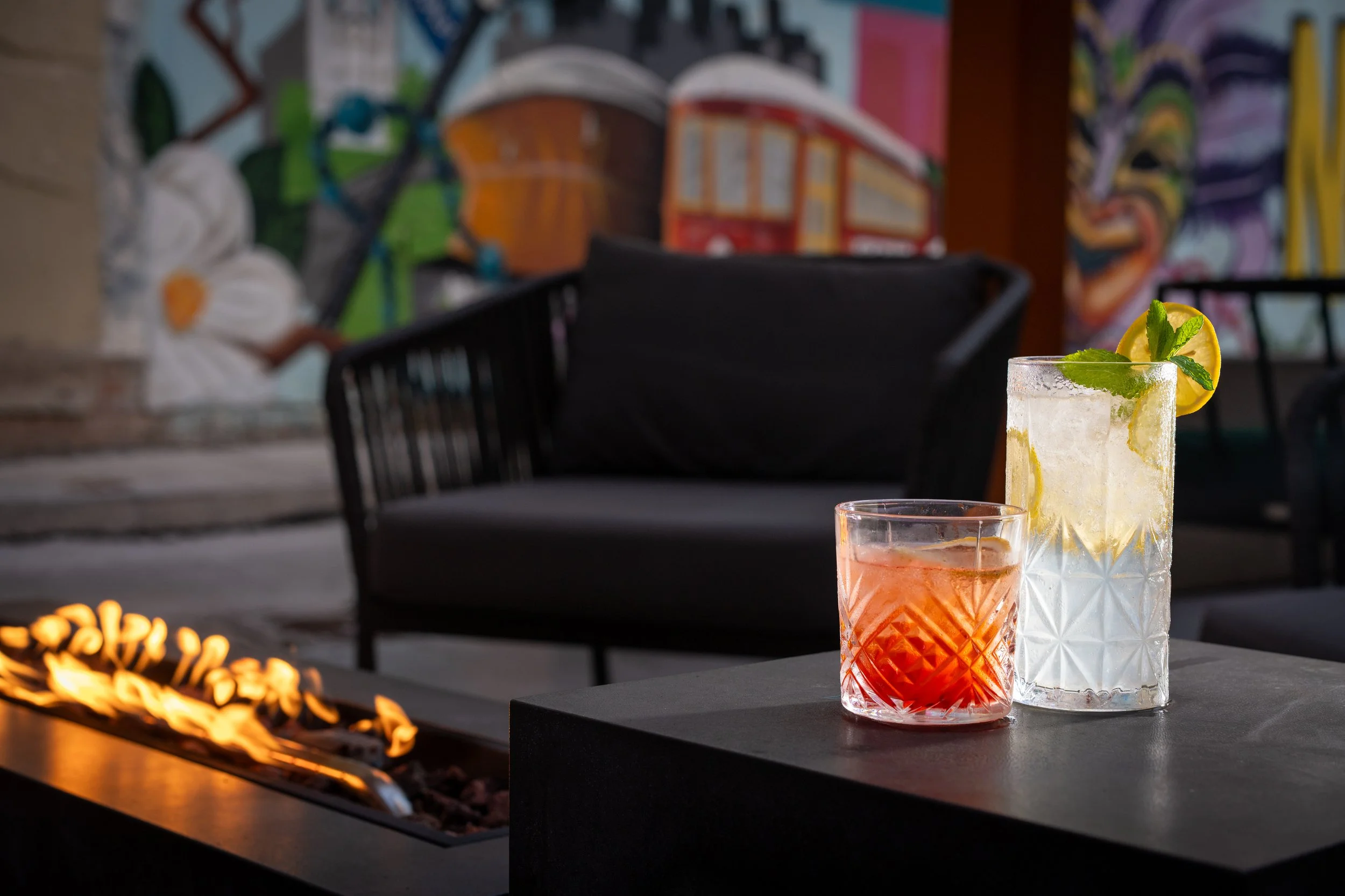 Two colorful drinks with lemon and mint garnishes on a black outdoor table in front of a graffiti-covered wall and a fire pit with glowing flames.