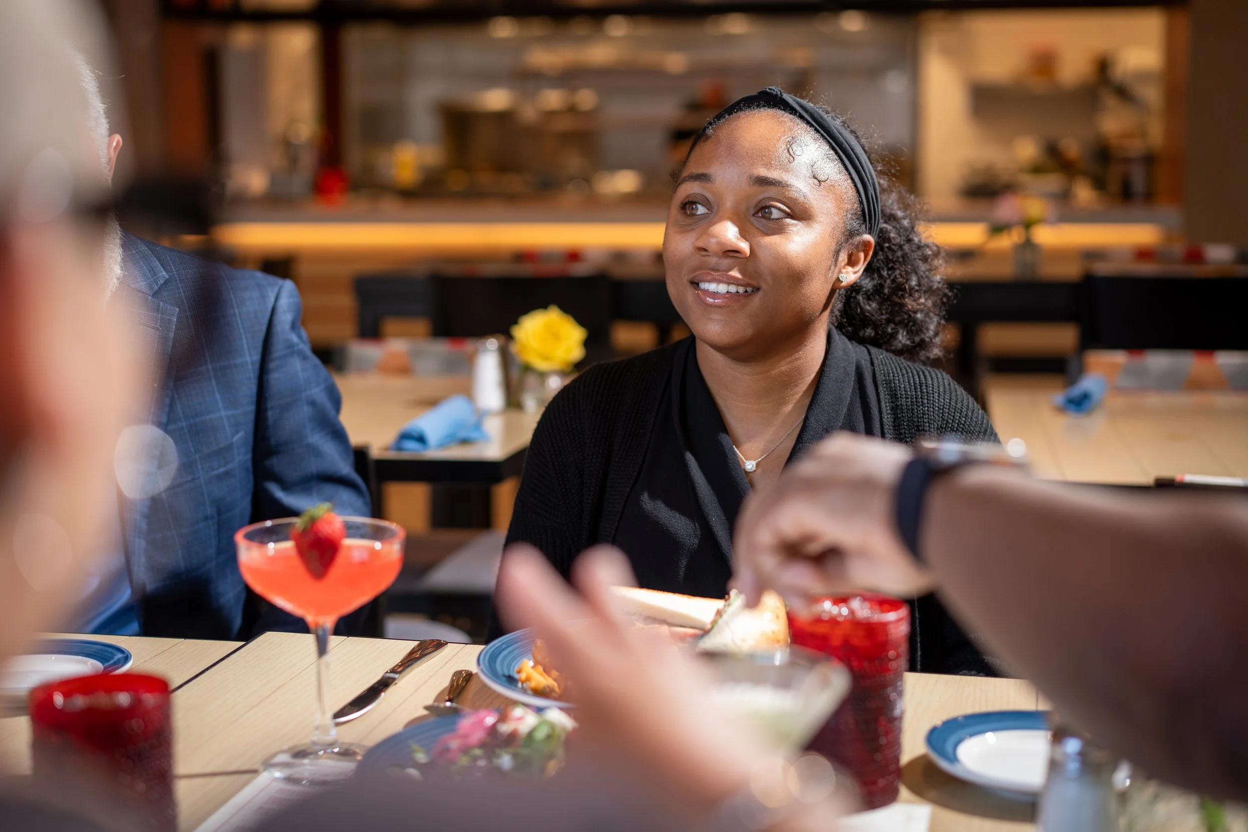 A woman with curly hair wearing a black headband and black top, smiling and looking to her right at a social gathering in a restaurant. She is sitting at a table with a colorful cocktail in a glass topped with a strawberry, and there are plates and utensils on the table.