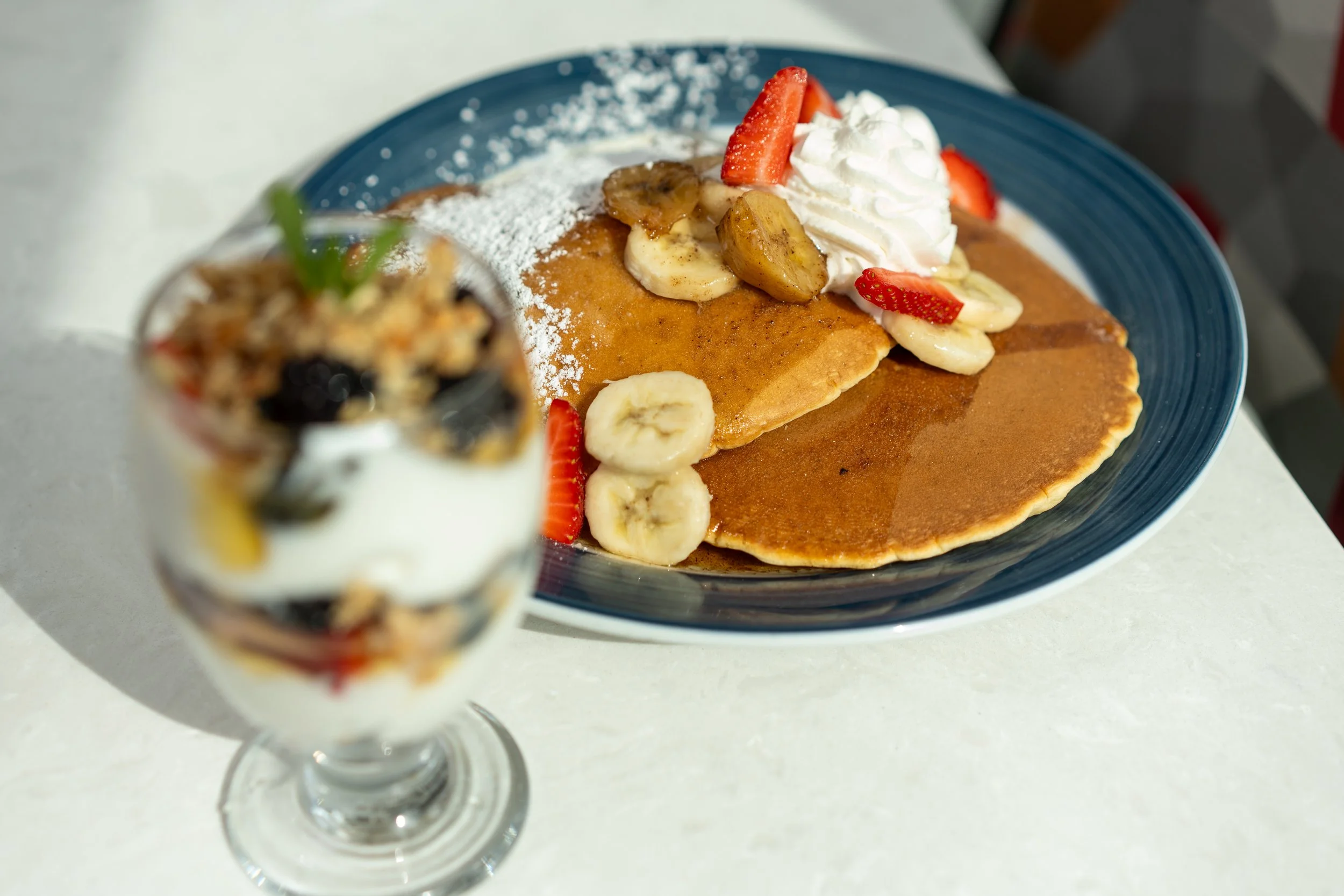 A plate of pancakes topped with bananas, strawberries, whipped cream, and powdered sugar, with a blurry dessert parfait in the foreground.