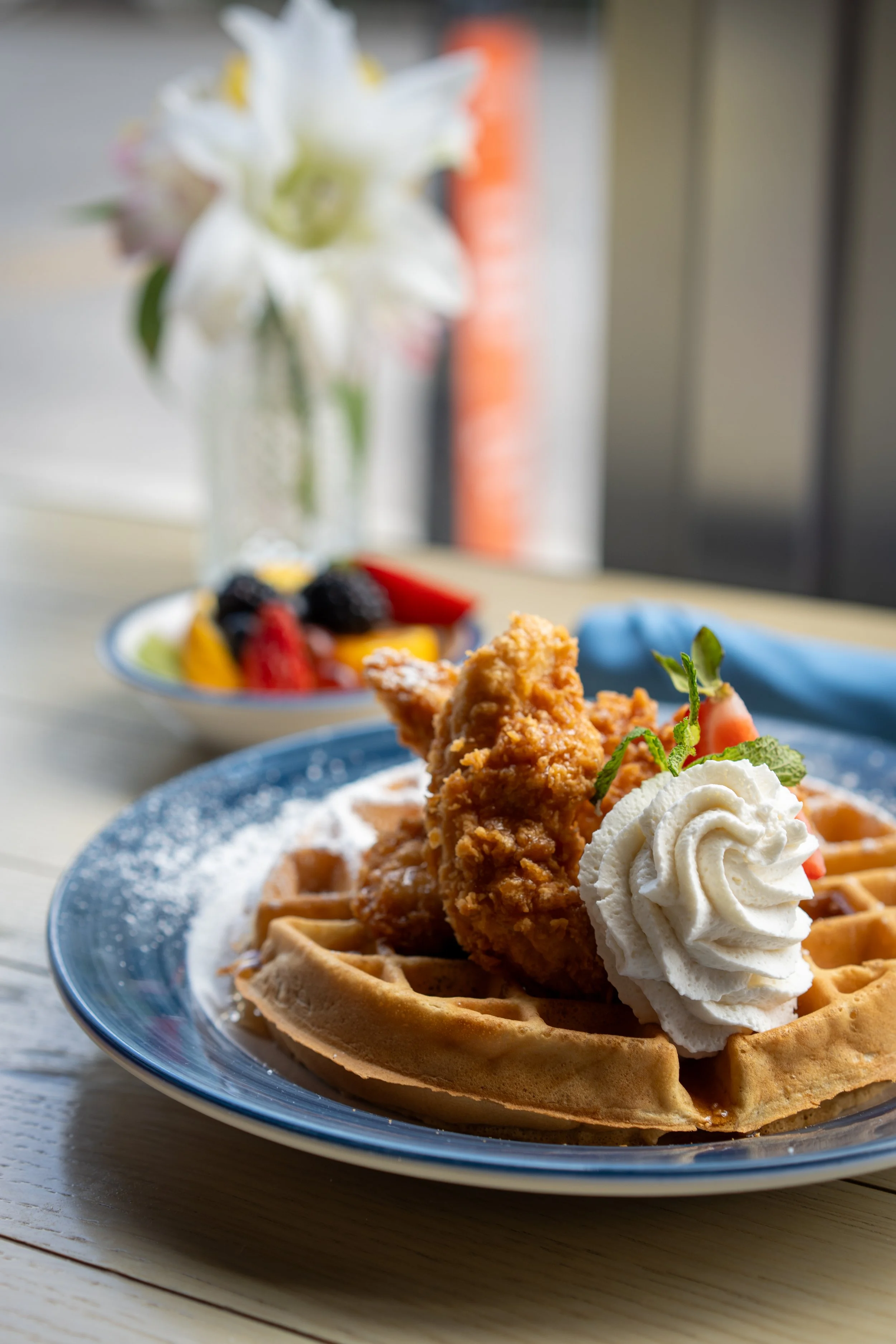 A waffle topped with fried chicken, whipped cream, powdered sugar, and a cherry, served on a blue plate.