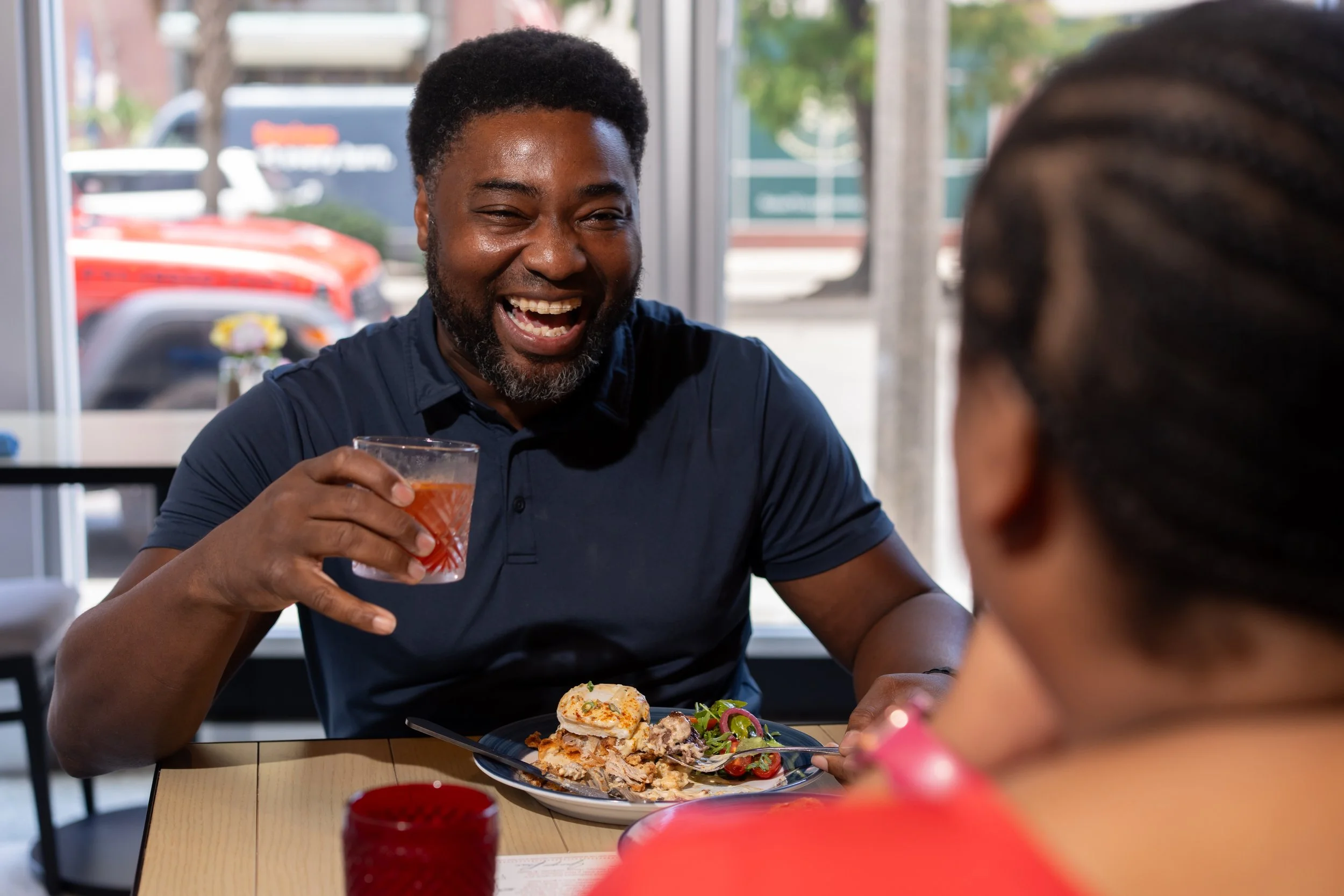 A man with a beard and short curly hair smiling as he holds a cocktail glass, sitting at a table with a plate of food, and talking to a woman with braided hair in a restaurant or cafe.