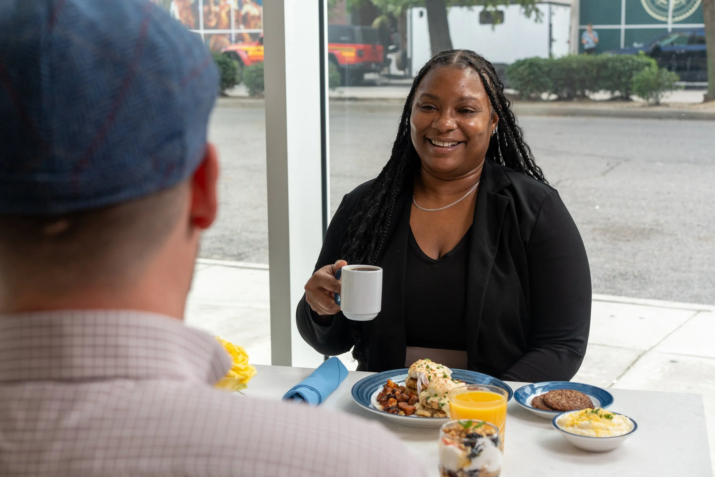 A woman with long dreadlocks smiling and holding a coffee mug, sitting at a table with various plate-sized foods, including mashed potatoes, gravy, and steak. A person wearing a blue cap and a checkered shirt is in the foreground, seen from behind. The setting appears to be a restaurant or dining area with large windows and outdoor scenery visible.