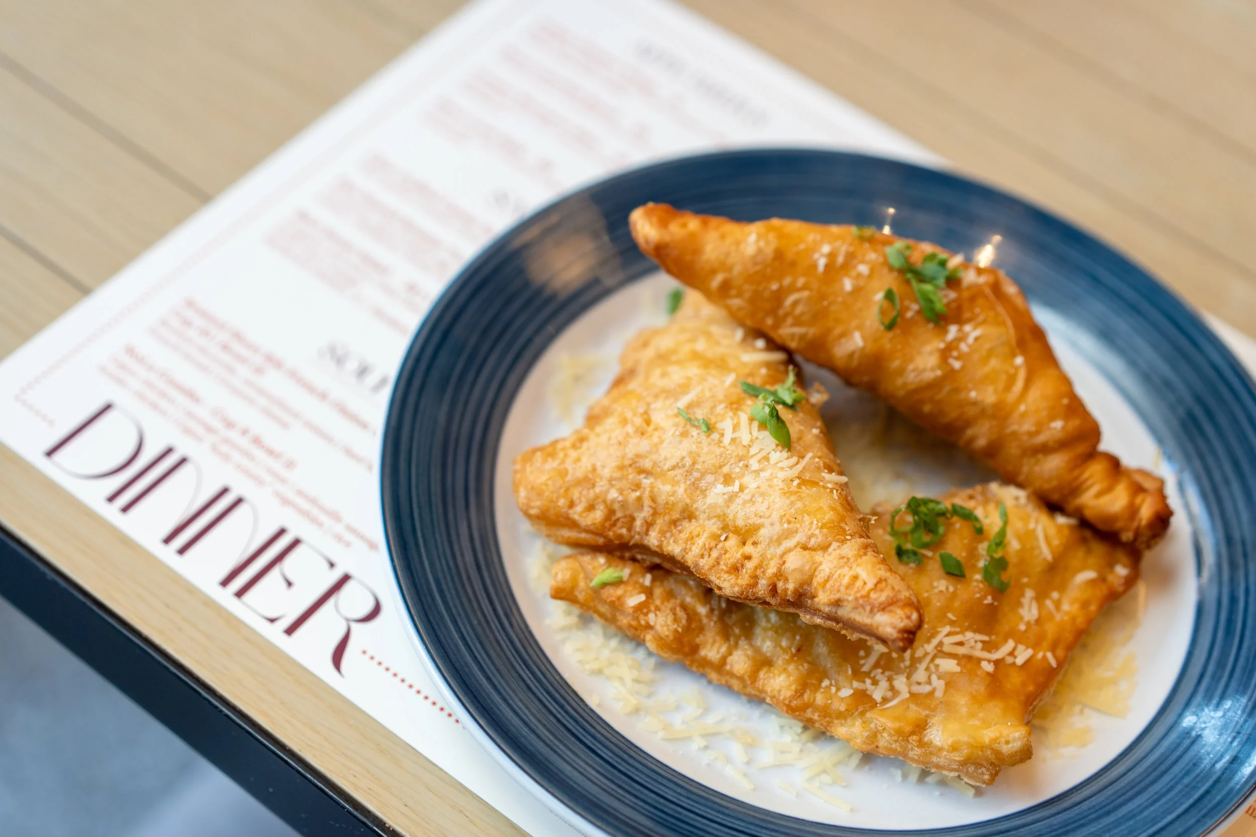 Two pieces of fried fish fillets topped with sauce and chopped green herbs, served on a blue plate with shredded cheese, resting on a wooden table with a printed menu underneath.