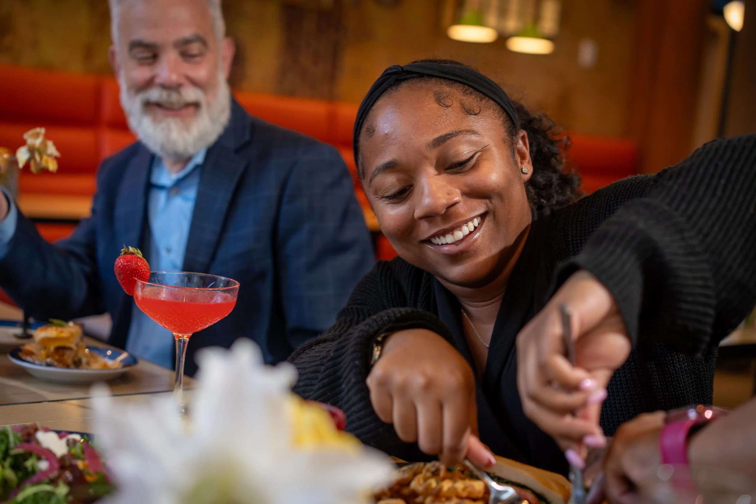 Two people, an older man and a young woman, are smiling and taking a selfie together at a restaurant table filled with various foods, drinks, and desserts. The man has a gray beard and is wearing a suit, while the woman is wearing a black headband and has her hair tied back.