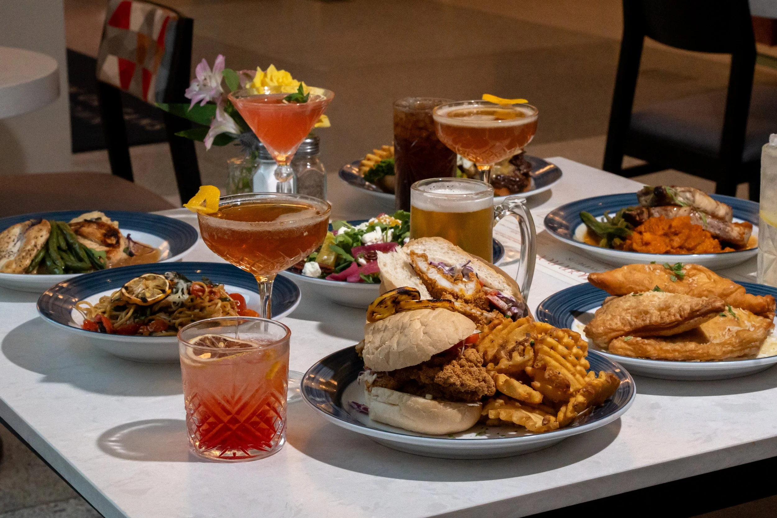 A table set with various dishes including sandwiches, fried chicken, pasta, and a salad, along with several drinks including beer and cocktails, at a dinner or party event.