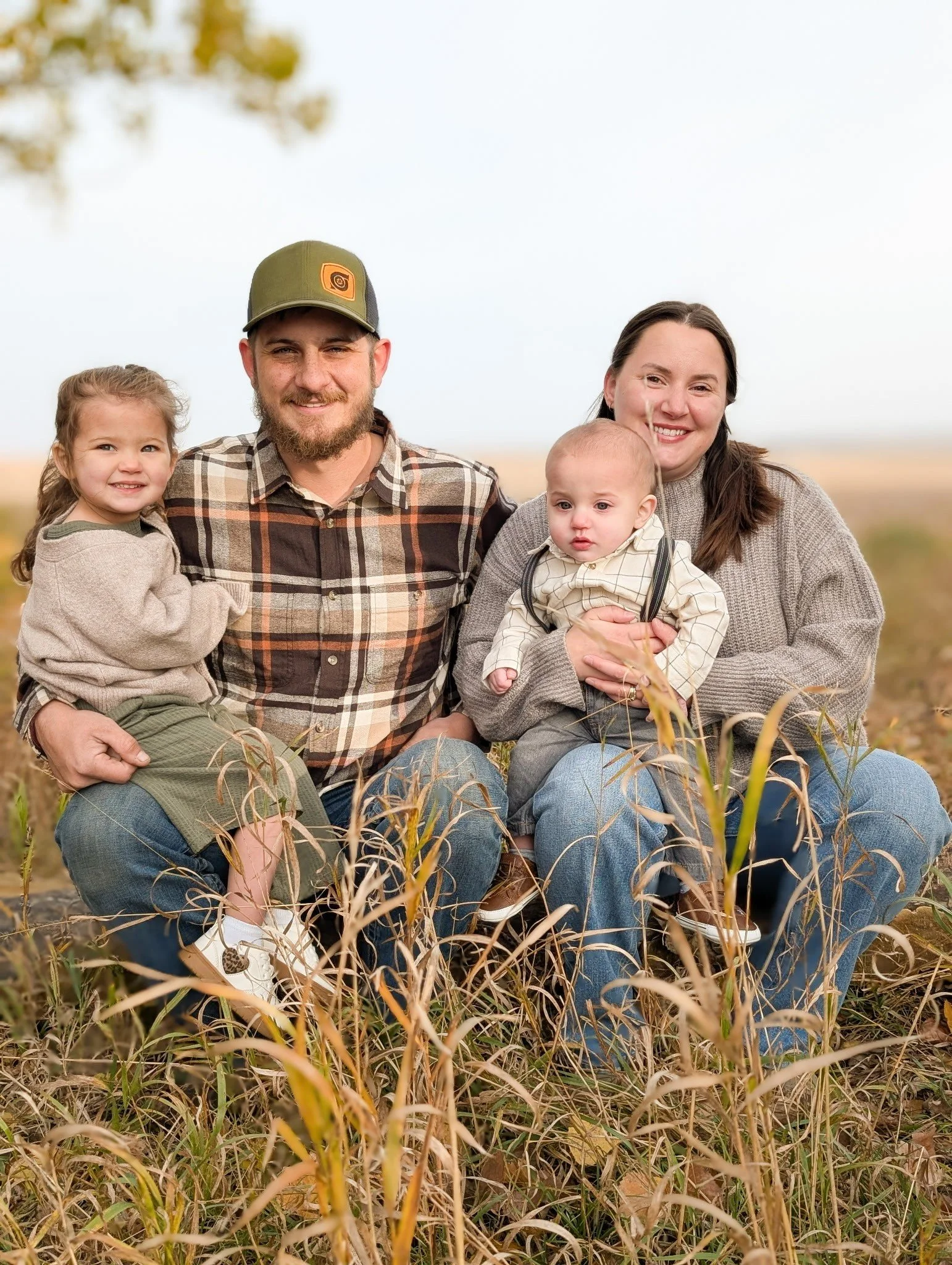 A family of four sitting in a field of tall grass outdoors, smiling at the camera. The family includes a young girl, a man with a beard and cap, a woman, and a baby. The sky is cloudy and there are some blurred trees in the background.