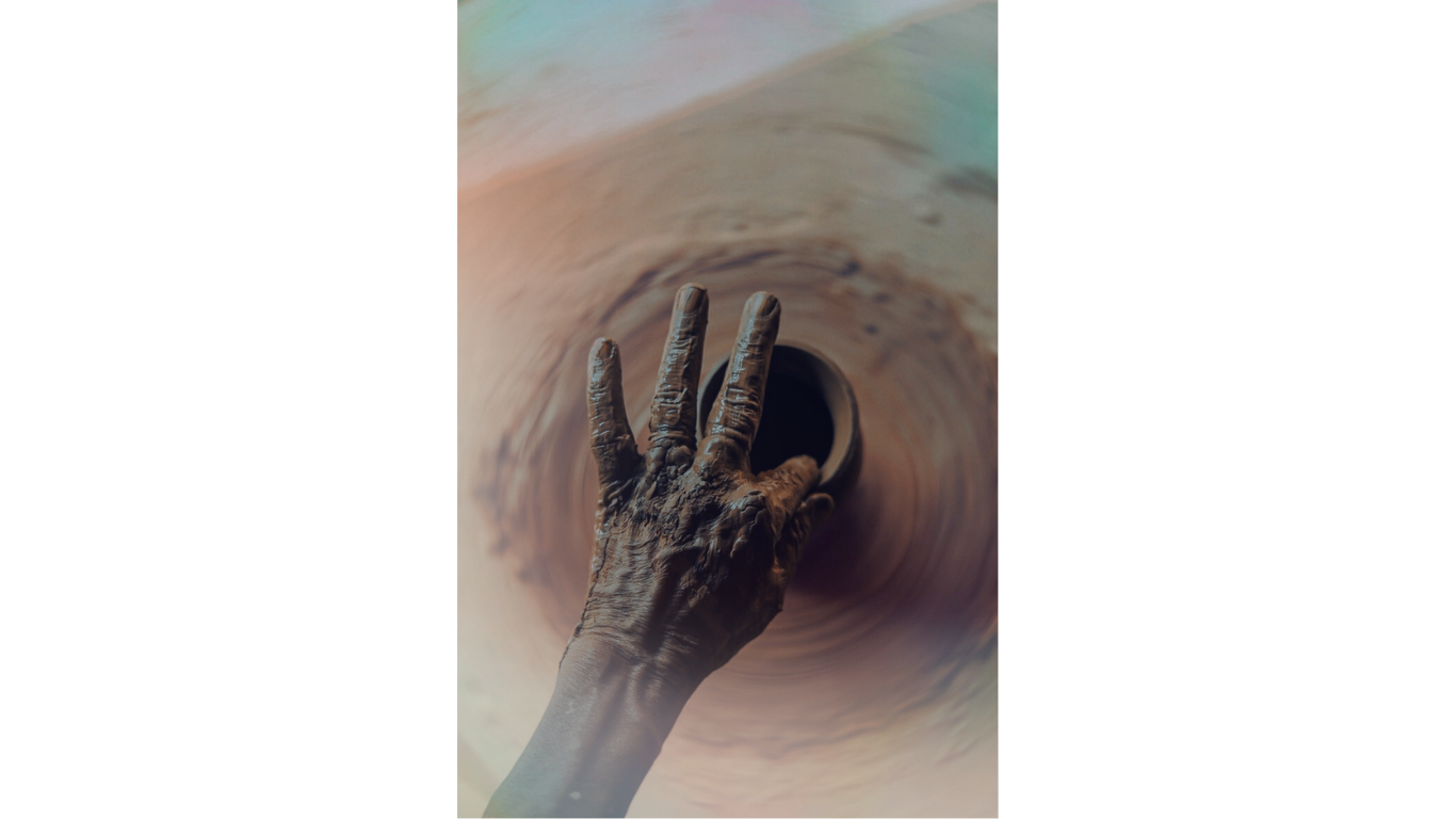 A person's hand covered in wet clay is reaching into a clay wheel or pottery wheel, shaping or centering the clay.