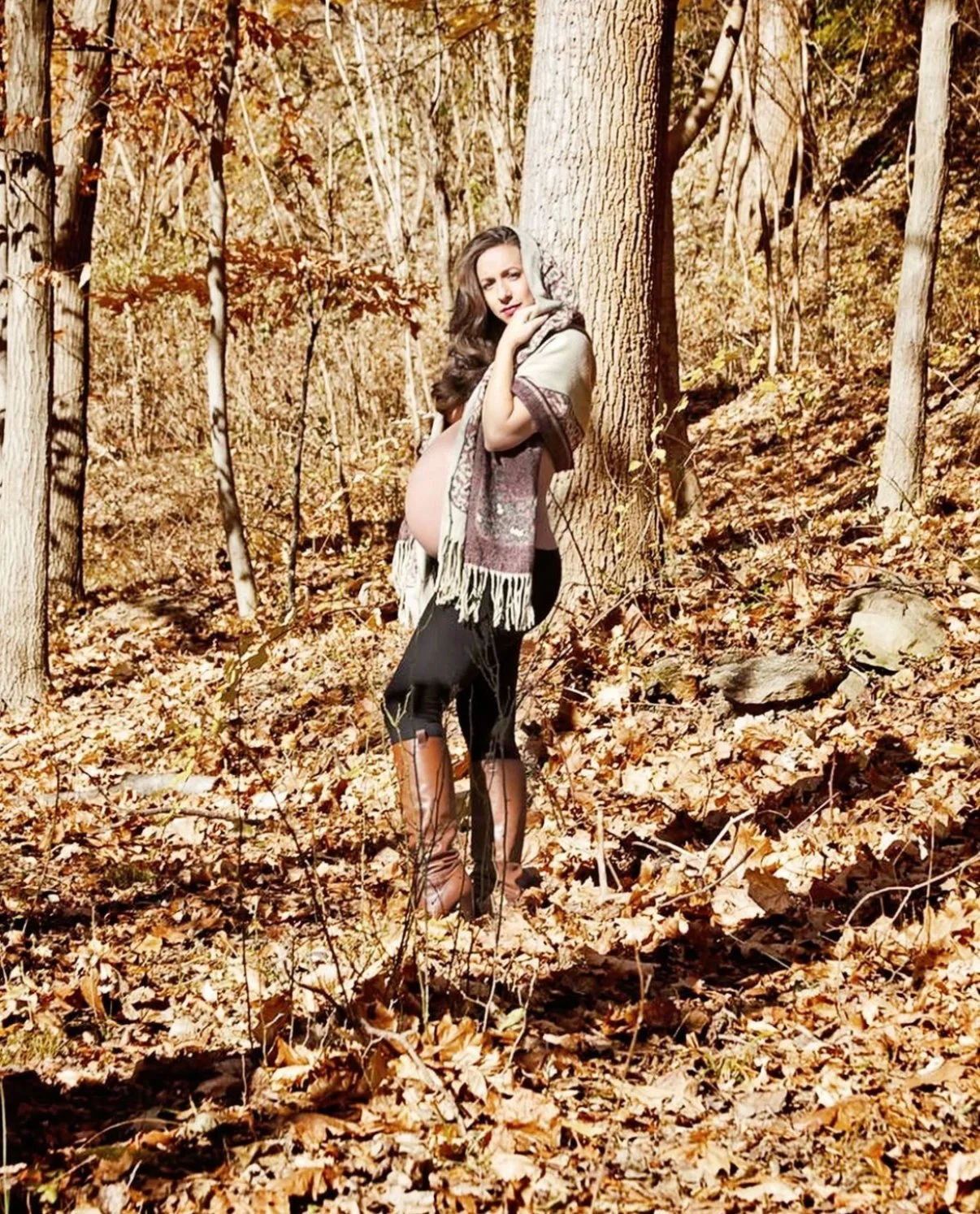 A woman standing in a forest with fall leaves on the ground, holding a scarf around her shoulders and heavily pregnant.