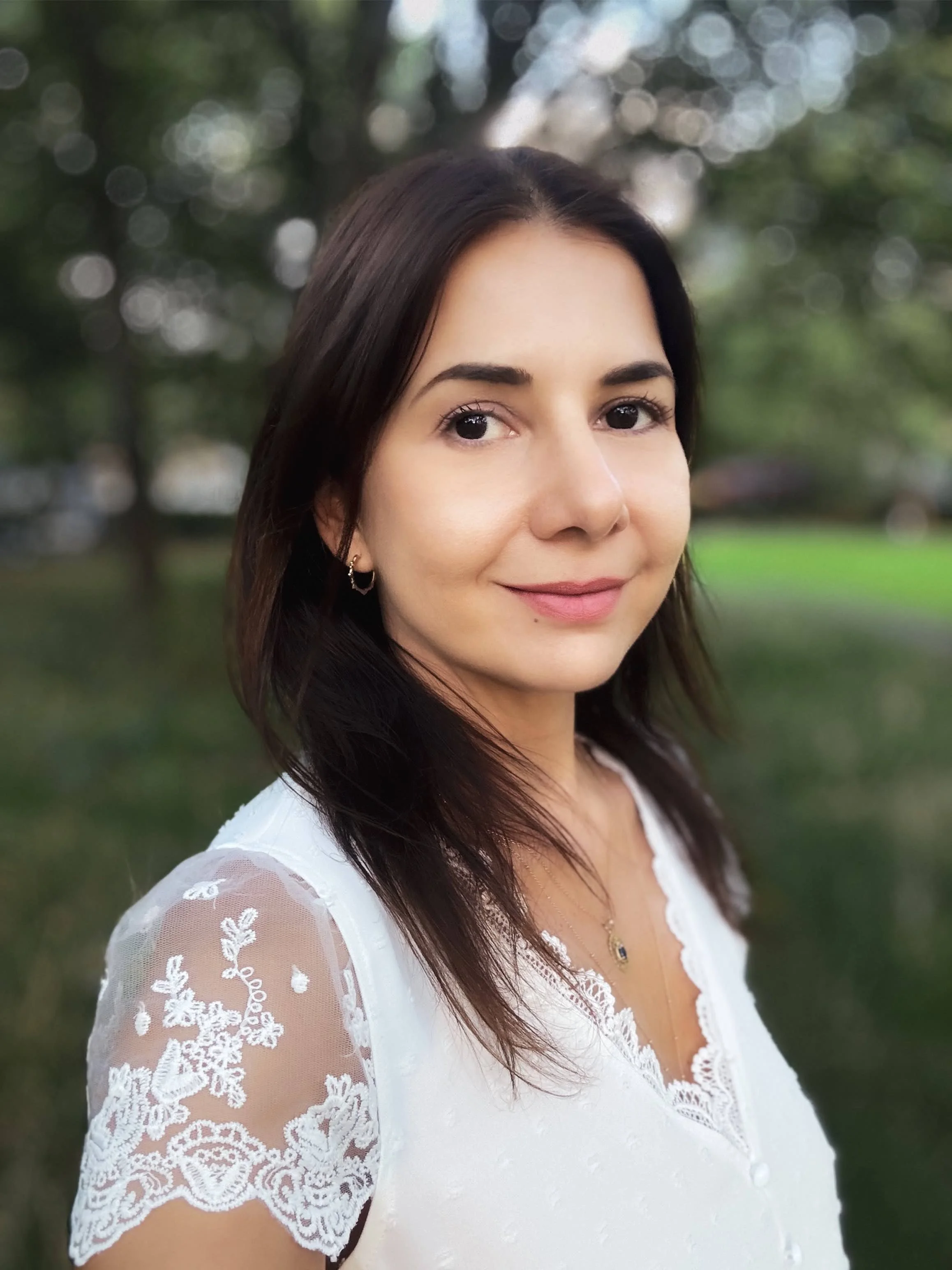 Portrait of a woman with dark brown hair, standing in front of greenery smiling