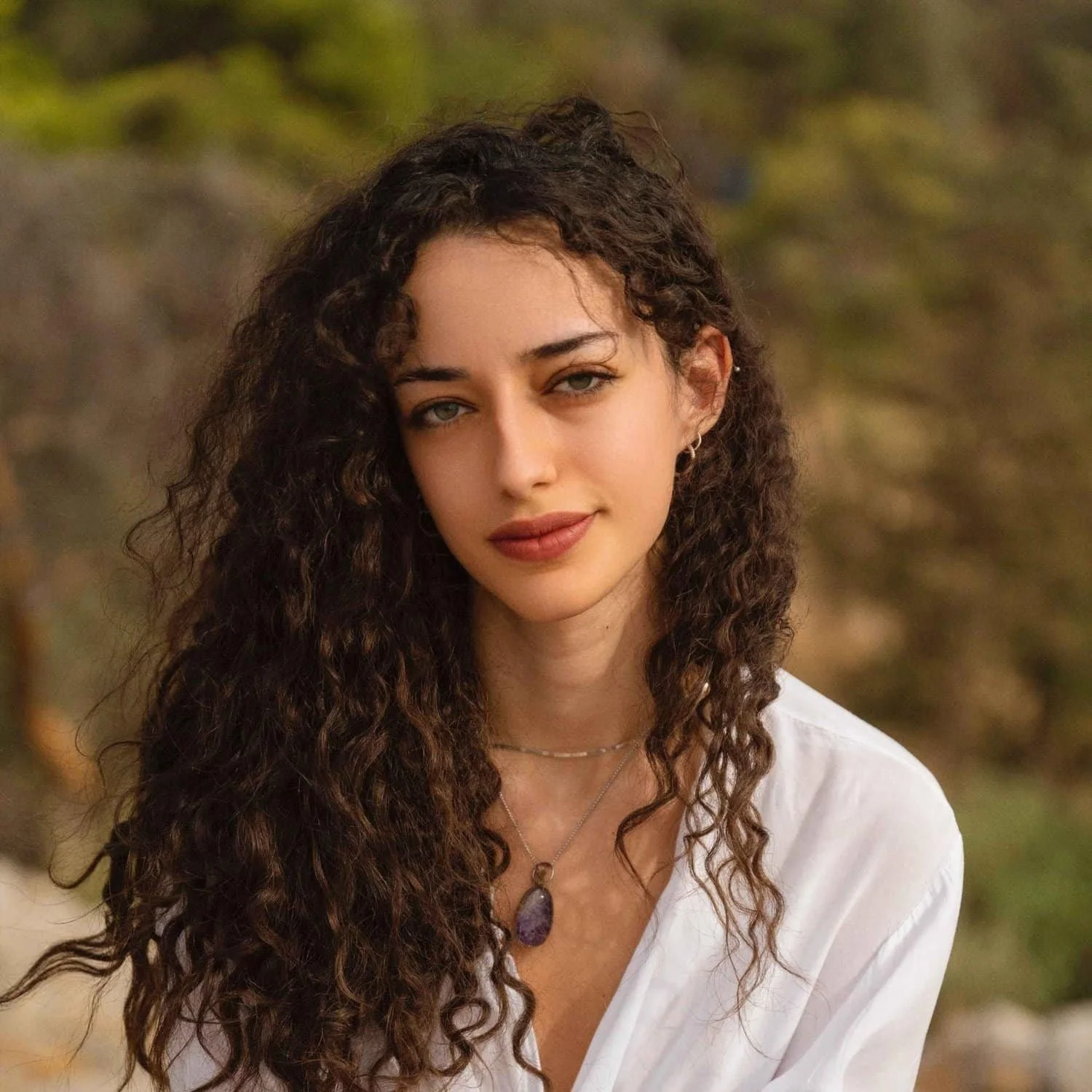 Portrait of a woman with curly dark brown hair, wearing a white top and white skirt, smiling while seated on a bench in front of a green in front of a background with green leaves