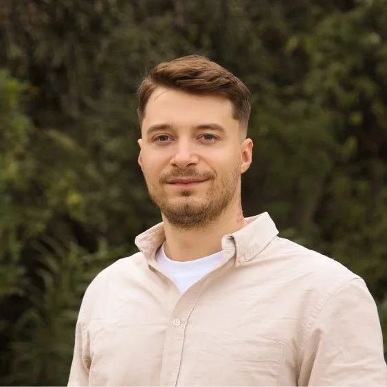 Portrait of a man with brown hair, wearing a cream shirt, standing in front of greenery smiling