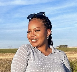 Portrait of a woman with braids, wearing a blue shirt and sunglasses above her head, standing in front of the blue sky smiling