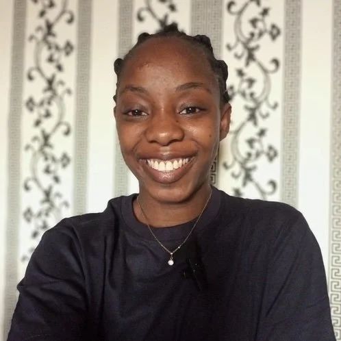 Portrait of a woman with cornrows, standing in front of a floral wallpaper smiling