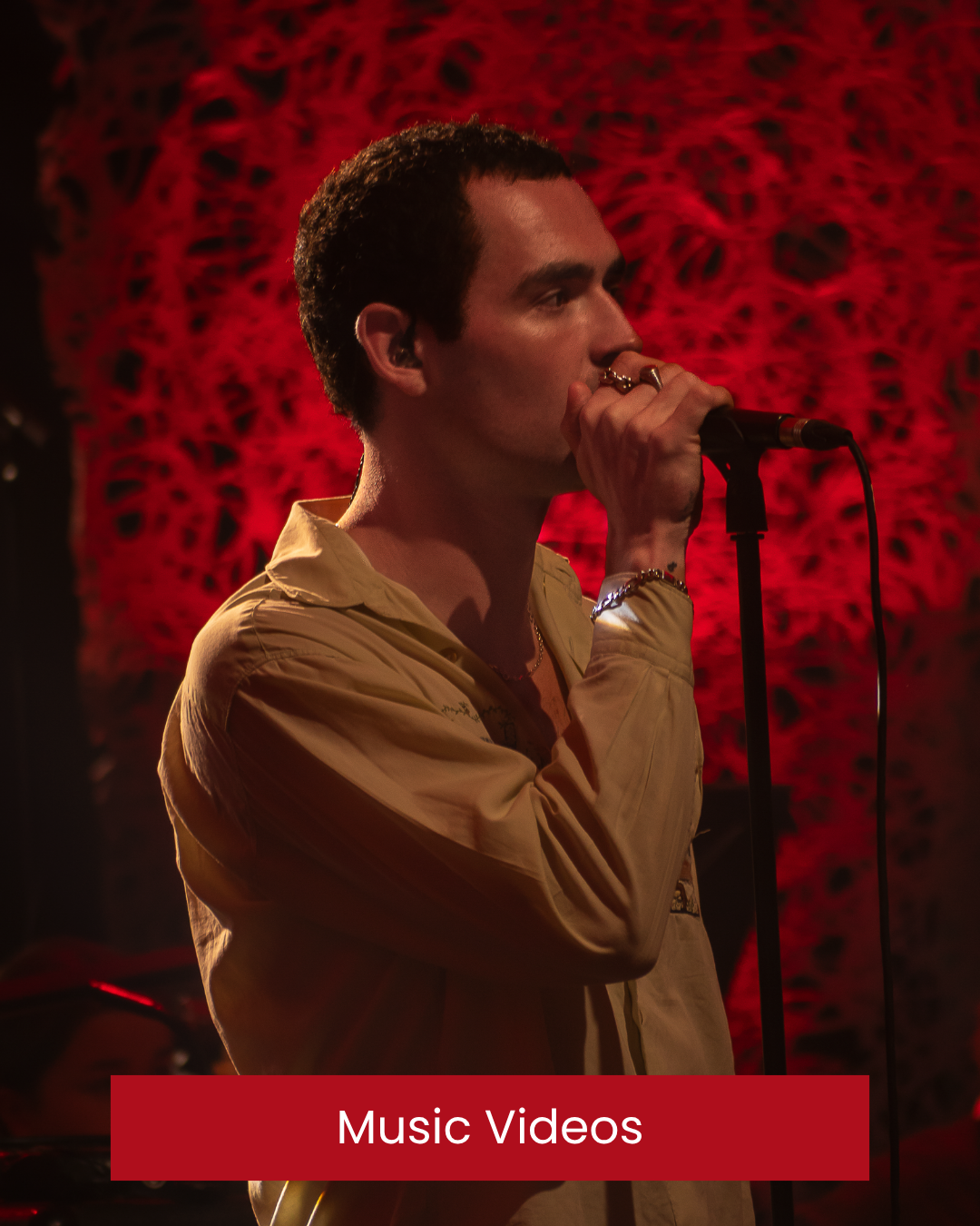 A male singer with dark, short hair wearing a yellow jacket, holding a microphone and performing on stage with a red, abstract backdrop. Murder Capital lead singer. RTE Culture Night.