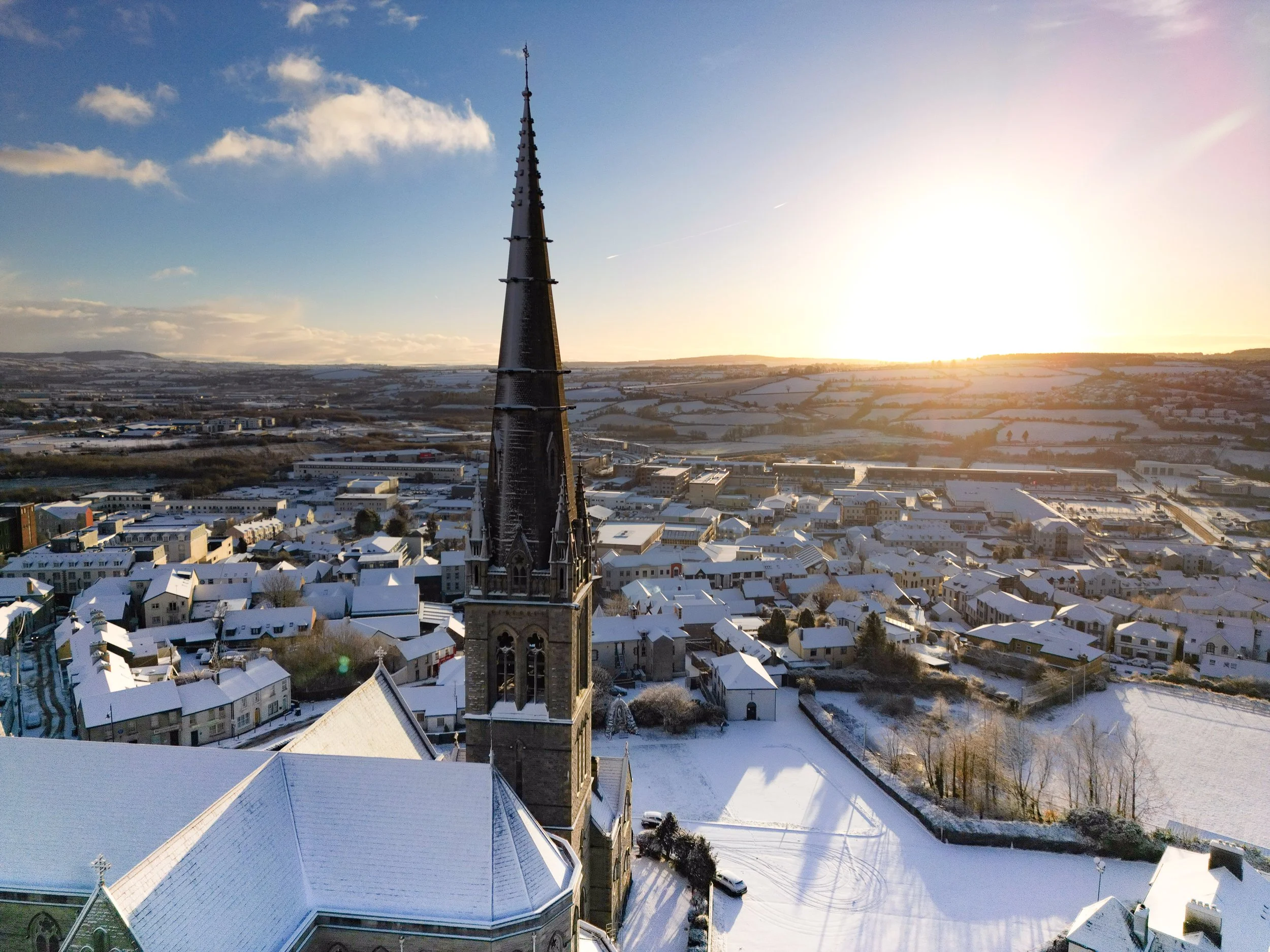 Aerial view of a town covered in snow with a church tower in the foreground, church's spire reaching into a partly cloudy sky during sunset.  Winter in Donegal. Winter in Ireland. Ireland Snow Photography. Saint Eunans Cathedral. Letterkenny. 