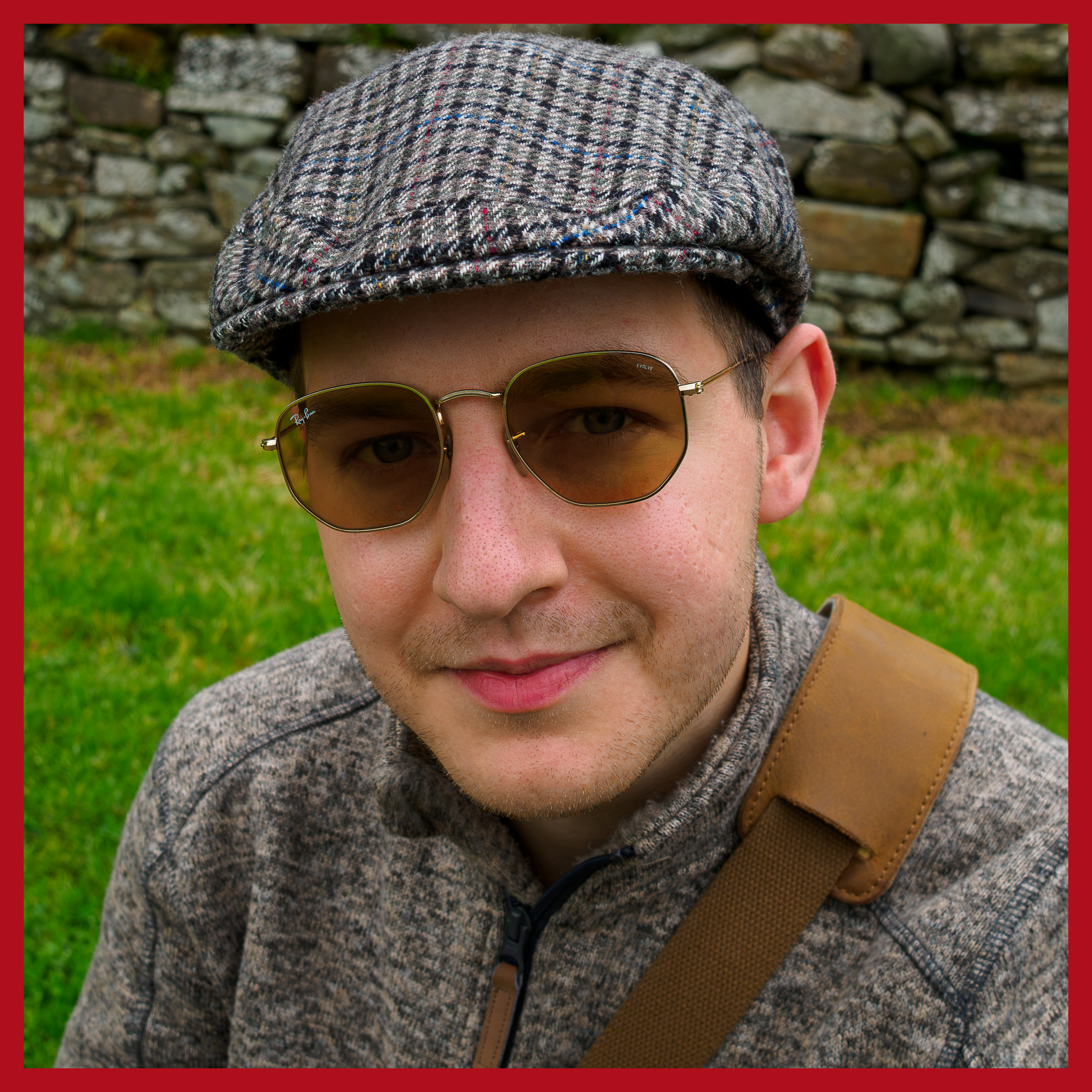 Close-up of a person wearing a gray tweed flat cap, sunglasses, and a gray hoodie, with a stone wall in the background. Artur Szotek is a Filmmaker based in Donegal. Expert in documentary films, marketing and social media content.