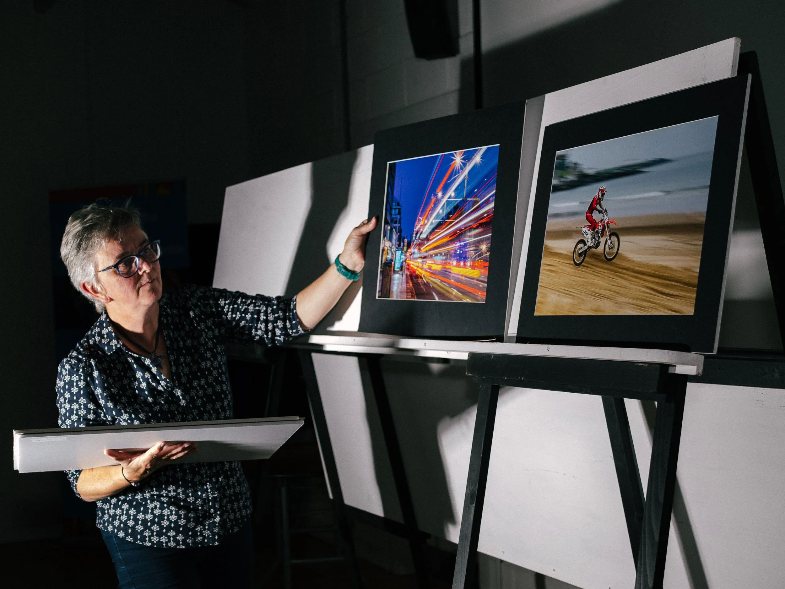 A woman with glasses and a patterned blouse looking at framed photographs on display at an art gallery or exhibition. Letterkenny Photographic Club. LKfoto. Interclub. Photography Club. 