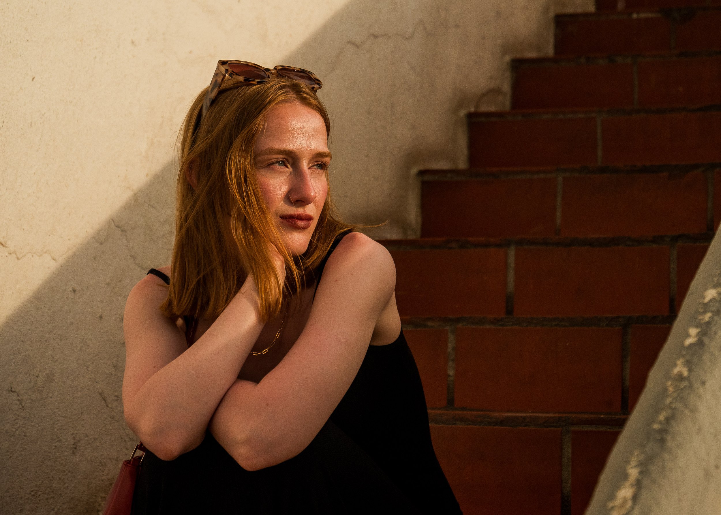 A woman with red hair, wearing glasses on her head, and a black top, sitting against a wall with stairs in the background. Marbella photoshoot. Female, clothing fashion lifestyle photoshoot. 