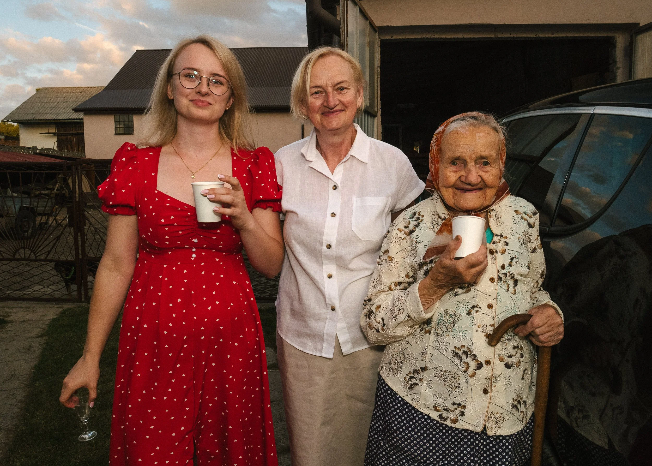 Three women standing together outdoors, holding cups, with a house and a car behind them, during the evening with a partly cloudy sky. three generations. family photography letterkenny donegal. 