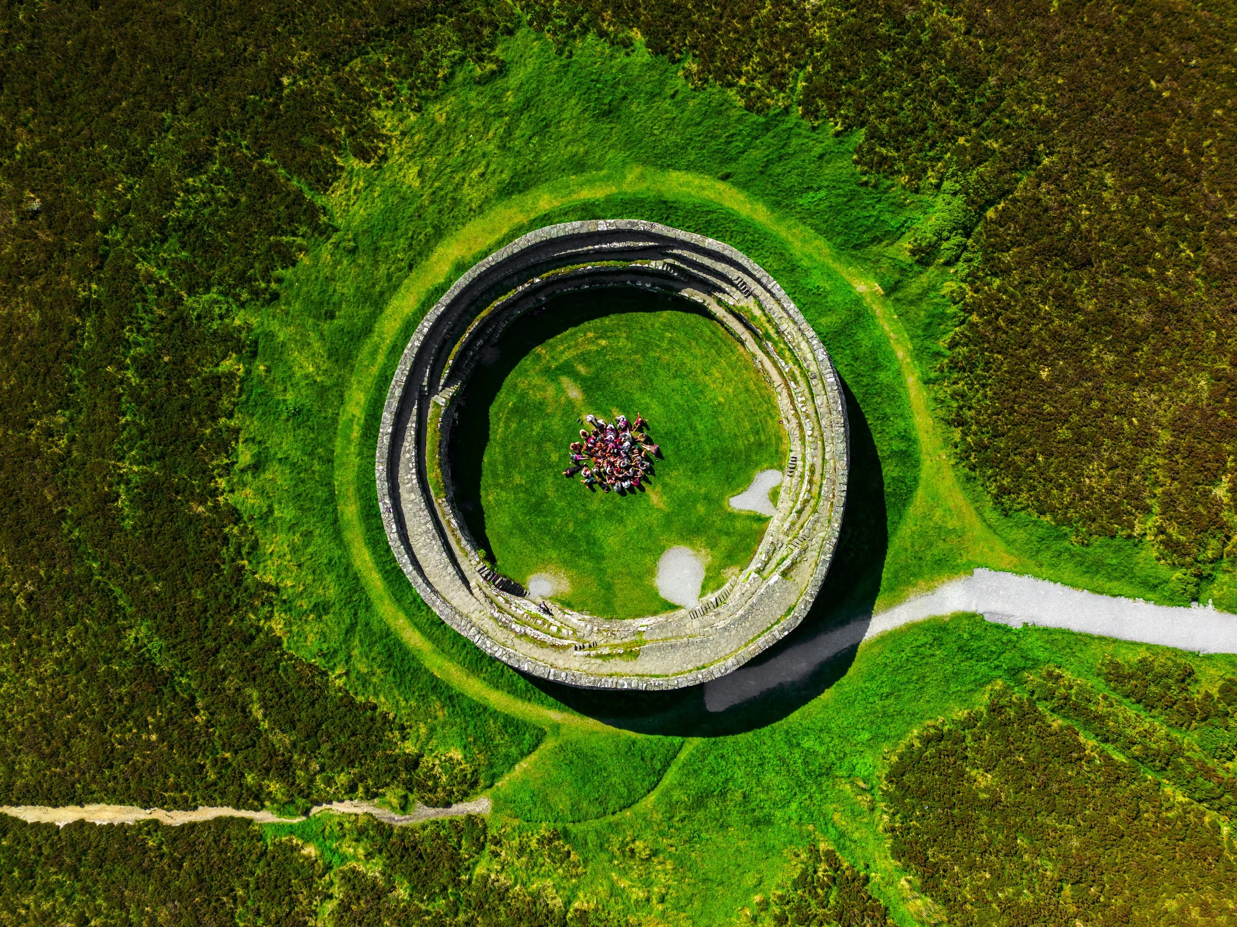 An aerial view of an old stone well with a group of people gathered at its base, surrounded by lush green grass and a dense forest. Grianán of Aileach. Greenan Mountain. Grianan of Aileach Hotel. Grianan Hotel. 