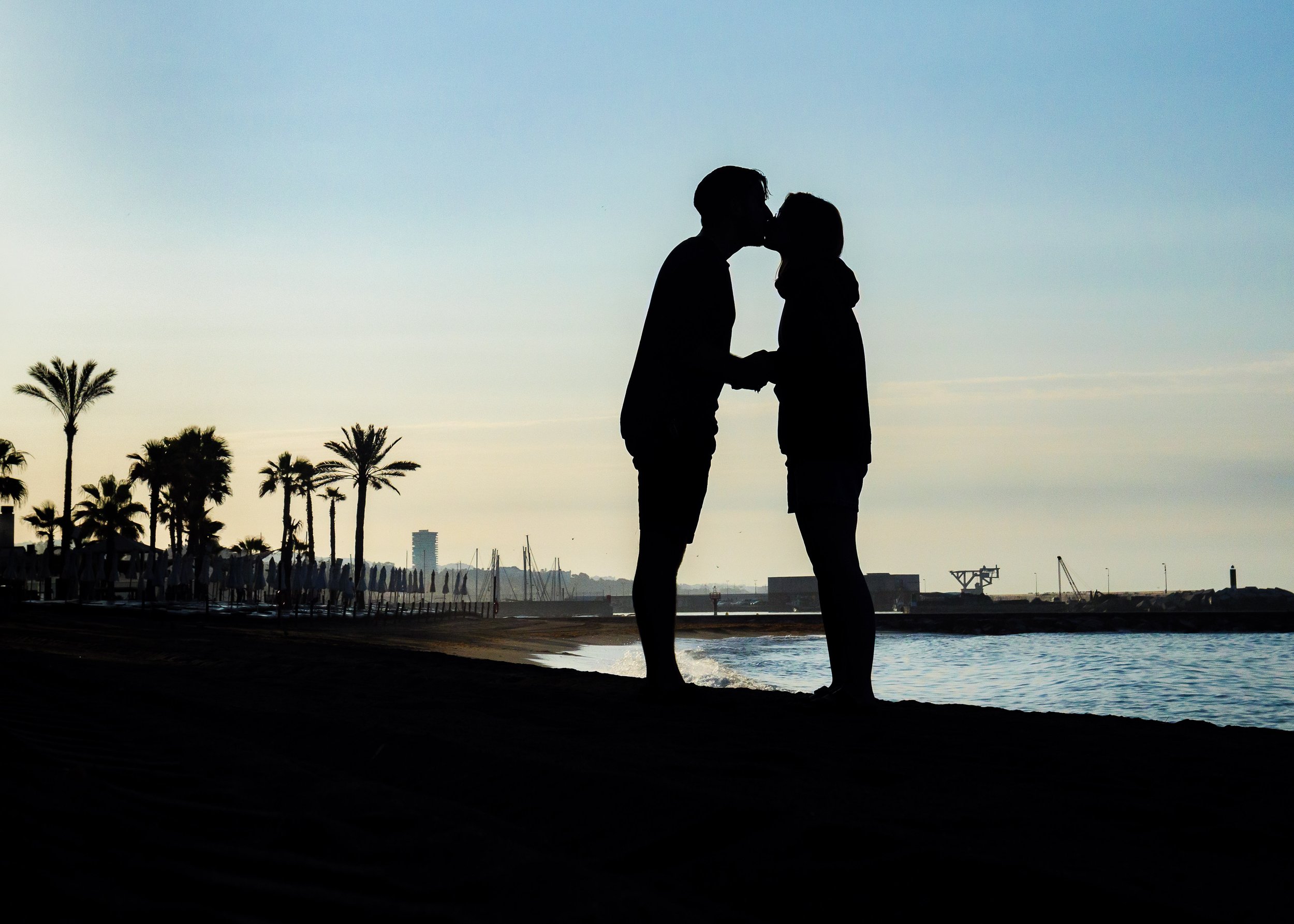 Silhouette of a couple kissing on a beach during sunset, with palm trees and harbor in the background. Marbella sunrise sunset couple photoshoot. Spain. Costa Brava. Engagement photography photoshoot. Wedding. Couple. 