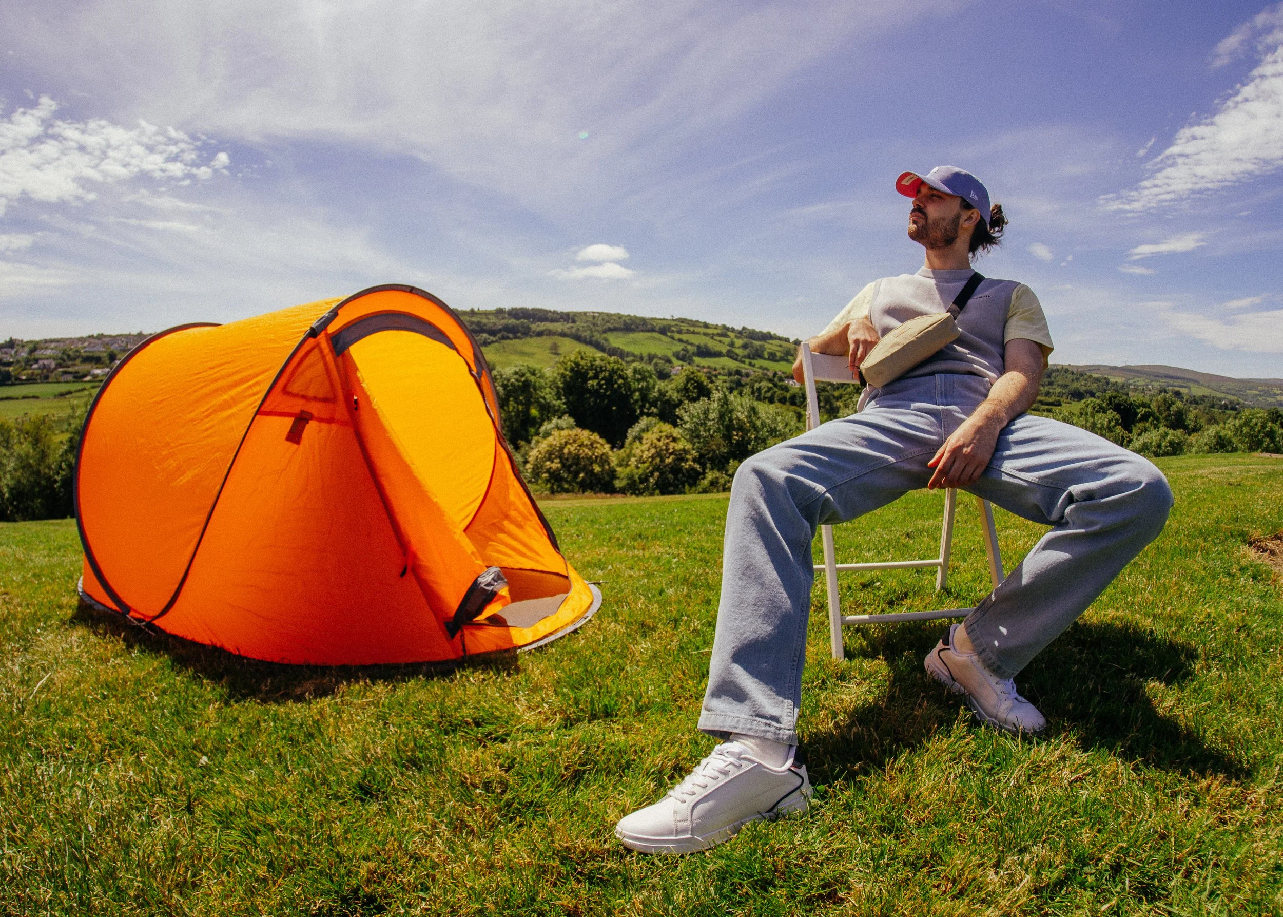 A man sitting on a lawn chair near an orange camping tent, with rolling green hills and a partly cloudy blue sky in the background. Edgar Malevic, model, e-commerce photoshoot in Donegal.  Evolve Clothing. Menswear. 