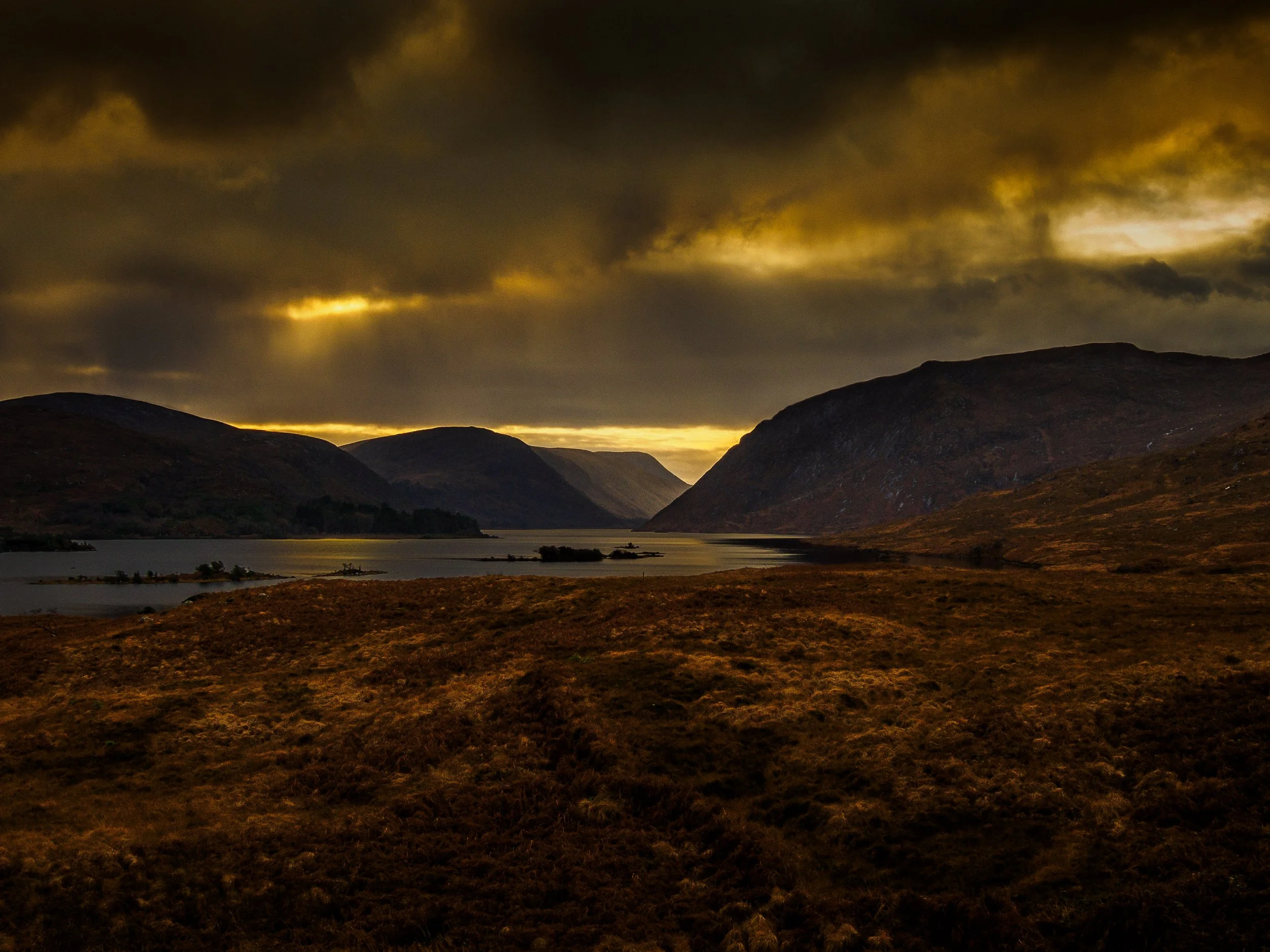 A landscape with dark, cloudy sky over rolling hills and a body of water, illuminated faintly by the setting or rising sun. Glenveagh National Park Sunset winter. 