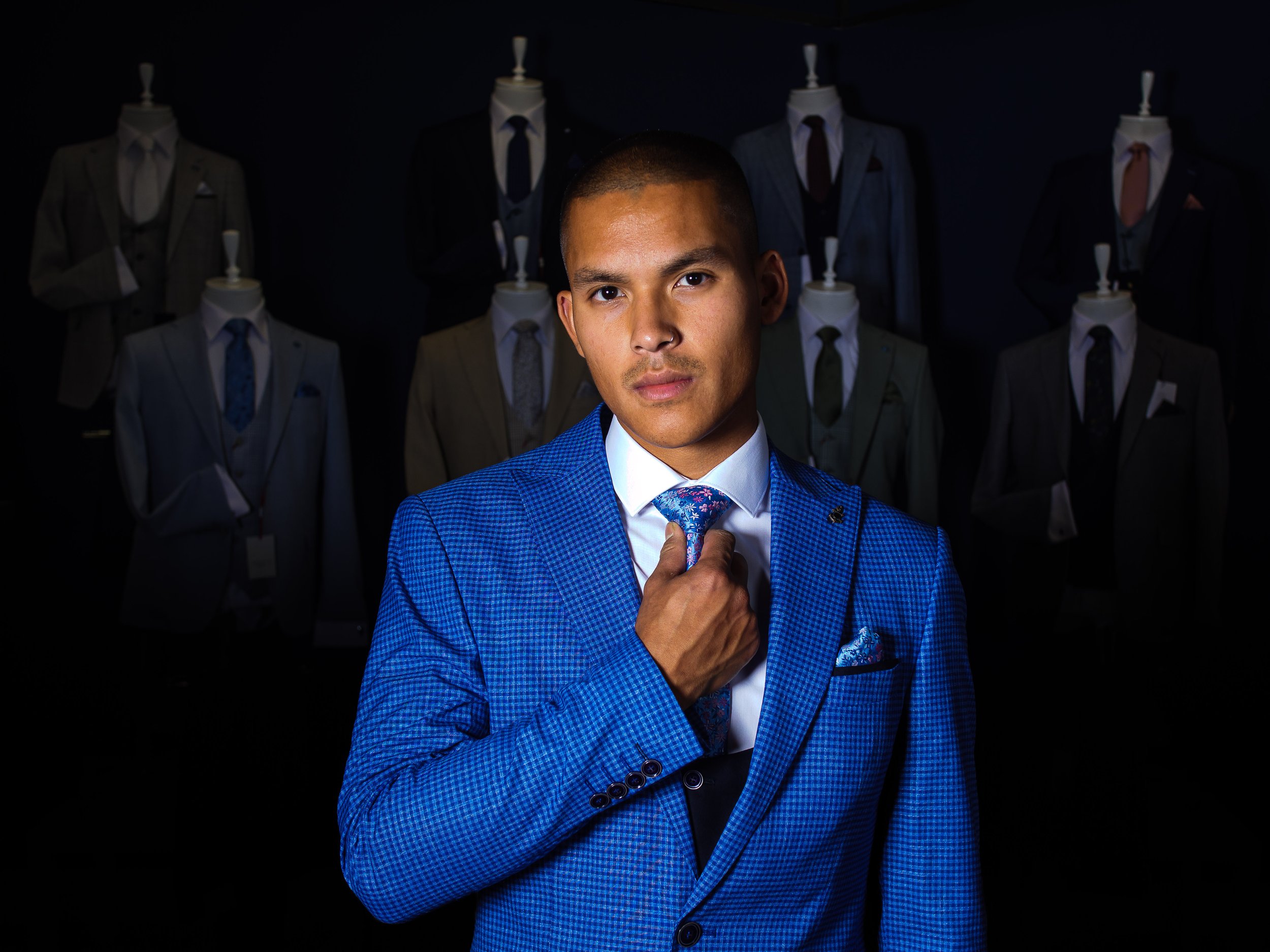 A young man in a blue checkered suit adjusting his tie, standing in front of mannequins dressed in various suits and ties, set against a dark background. Evolve clothing, model, e-commerce photoshoot in Donegal. Menswear.