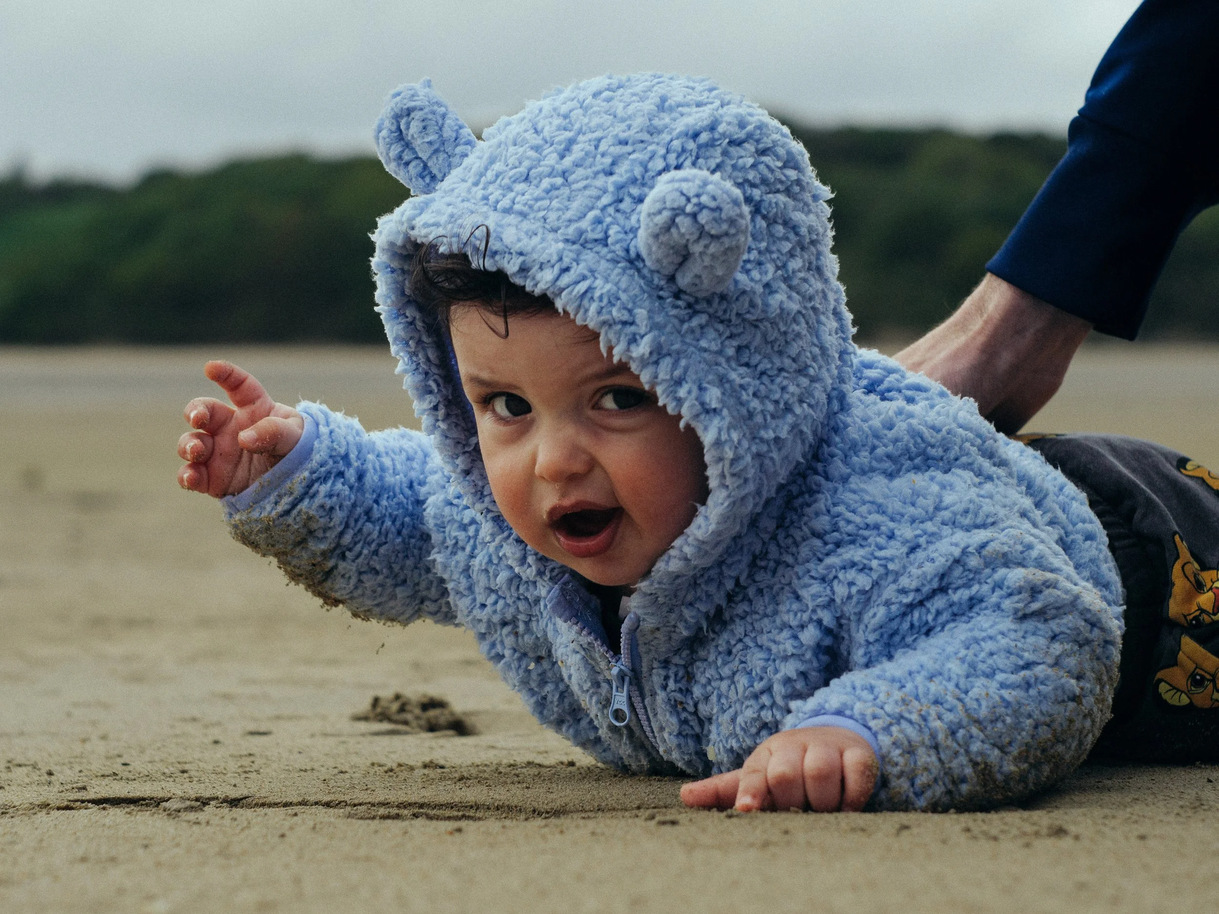 A young child crawling on sandy beach, wearing a light blue fluffy hoodie with bear ears, with sand on their fingers and face, and an adult's hand holding onto their hoodie. Newborn photoshoot Donegal. 