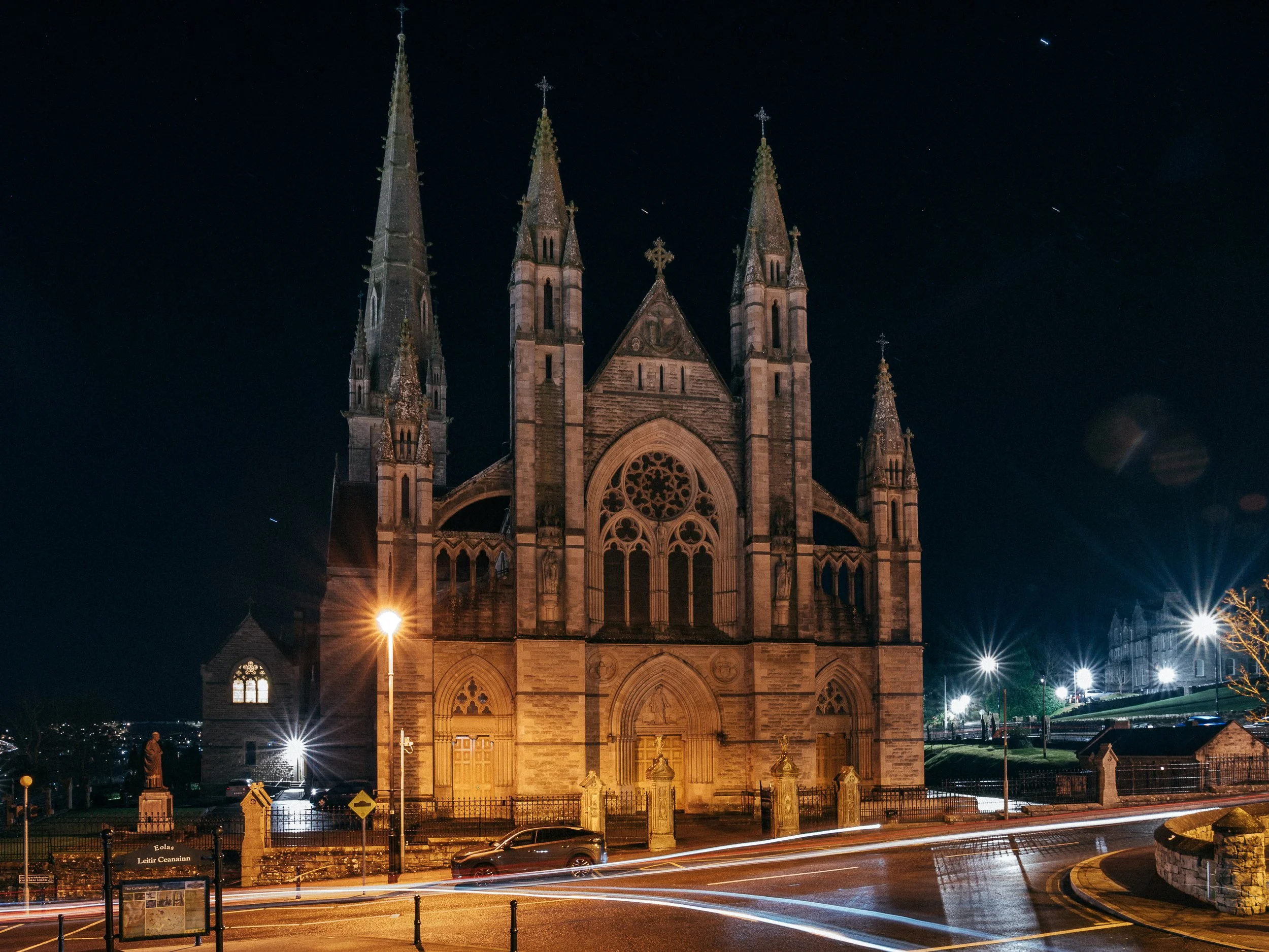 Night view of a large Gothic-style church with tall spires and ornate stained glass windows, illuminated against the night sky. Streetlights and moving vehicle lights create streaks of light at the street level. Letterkenny Saint Eunans Cathedral. 