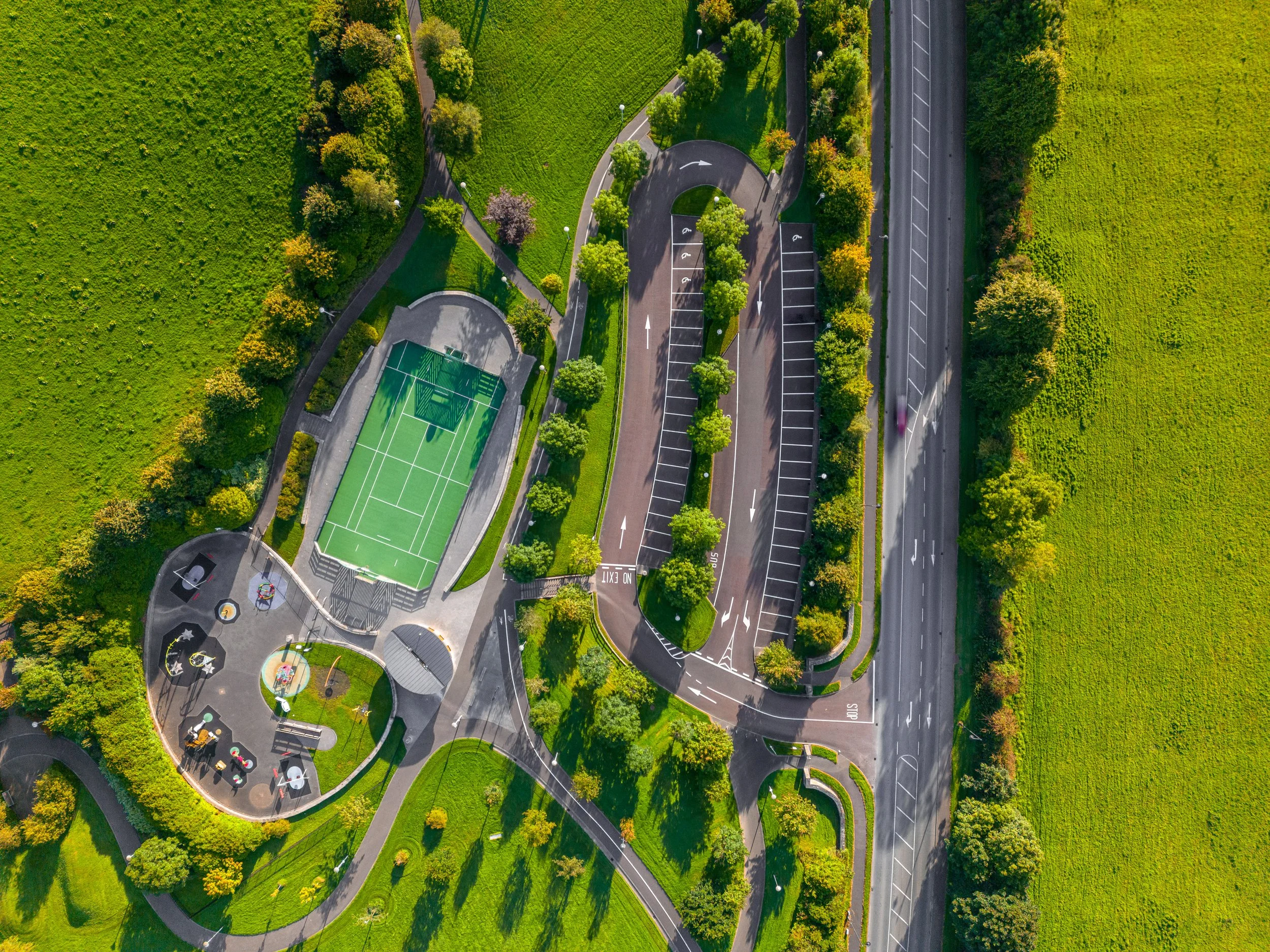 An aerial view of a park with walking paths, green lawns, trees, a parking lot, a basketball court, and a multi-ride playground. Ballymacool Park in Letterkenny Donegal Ireland. Drone videographer photographer in Donegal. 