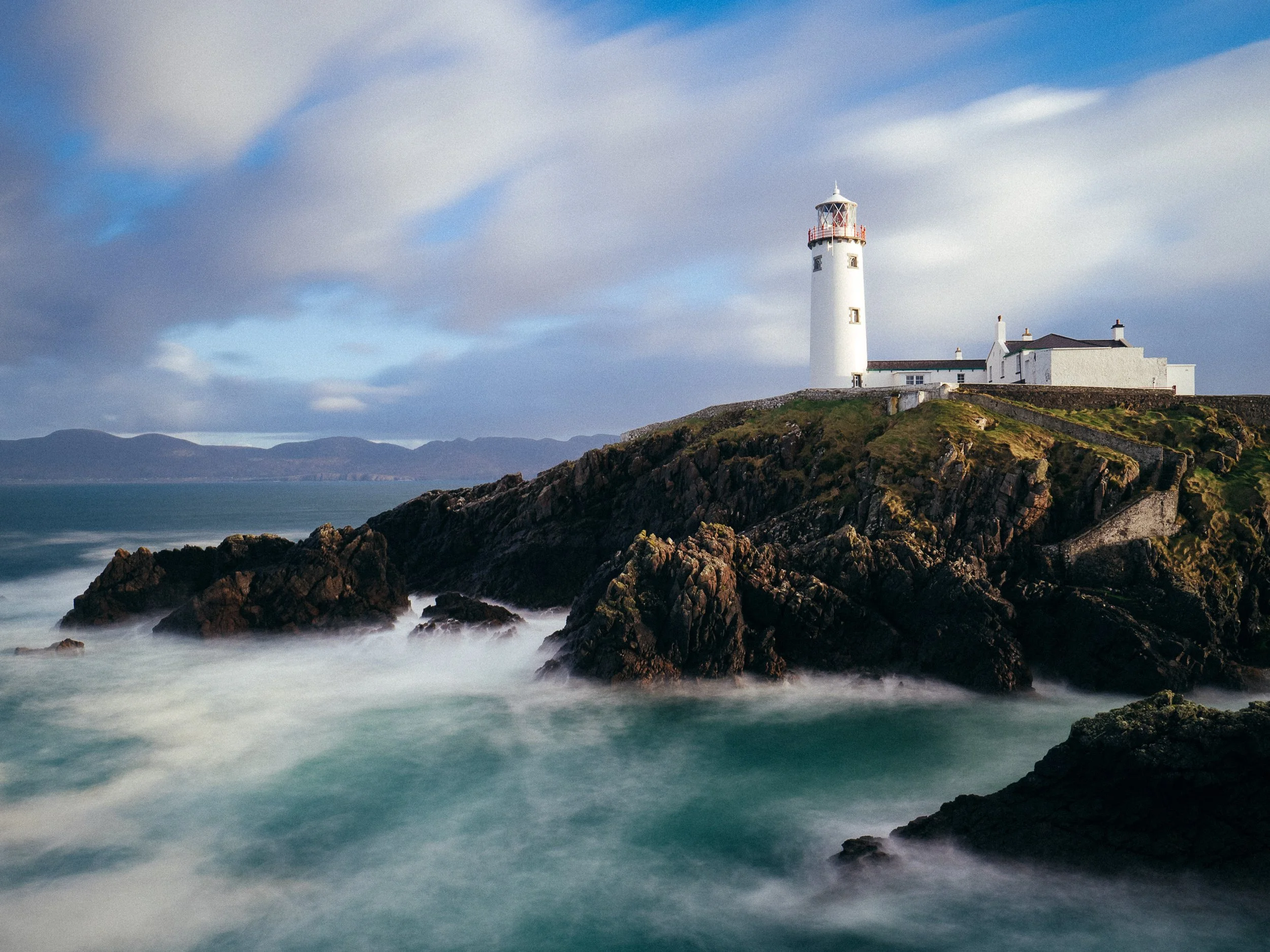 A lighthouse on a rocky coastline with ocean waves in the foreground and a cloudy sky in the background. Fanad Lighthouse. inishowen peninsula. Cliffs. Wild Atlantic Way. Visit Donegal. Visit Letterkenny. 