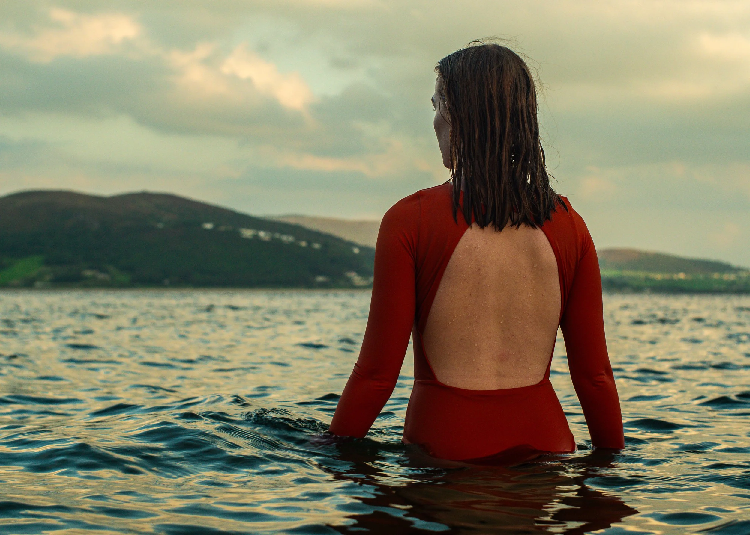 A woman with wet hair wearing a red backless dress standing in a body of water facing away with hills and cloudy sky in the background. Surfing in Donegal. Rathmullan beach. Kinnegar beach. Portsalon Beach. Buncrana beach. 