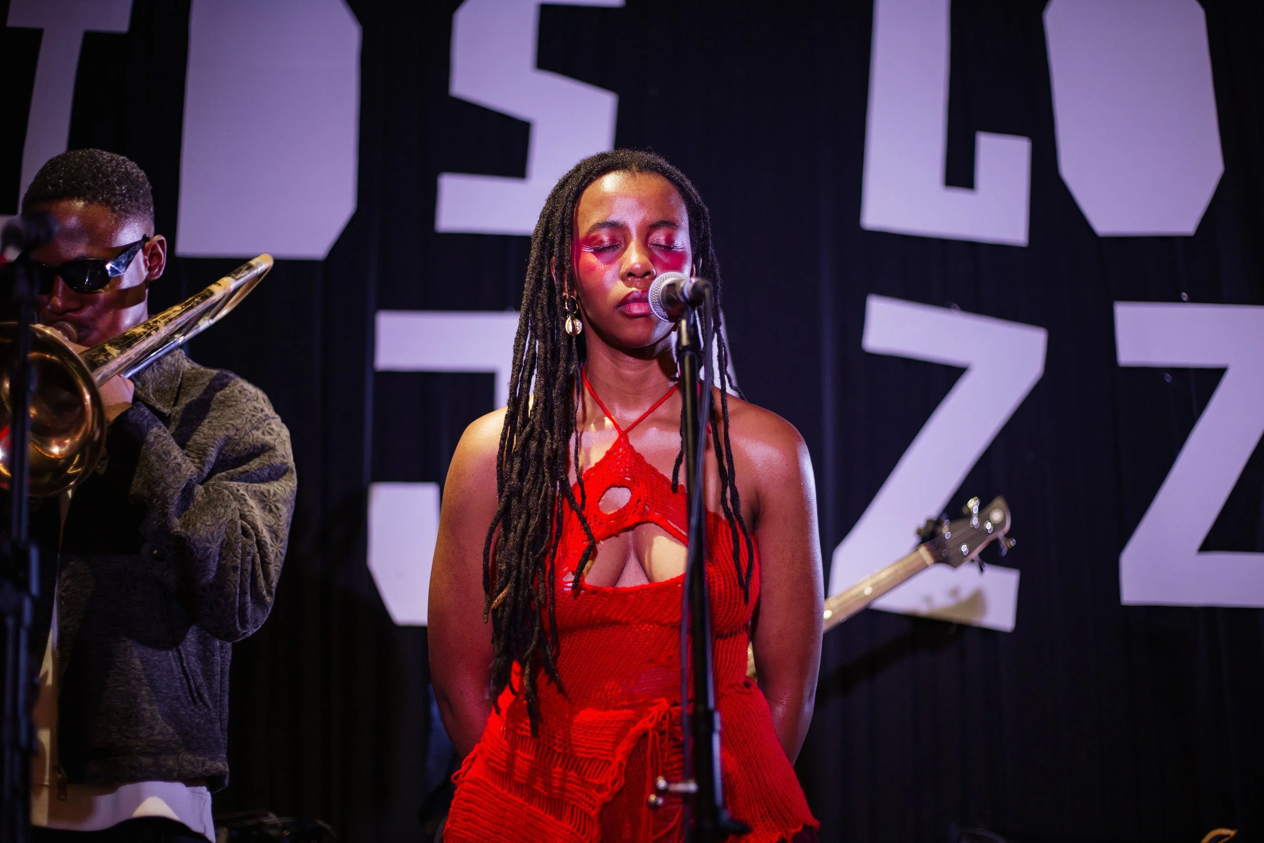 A woman with long dreadlocks, wearing a red crochet dress, singing with her eyes closed into a microphone on stage. A musician playing a trombone is visible on her left. Black stage backdrop with large white letters.