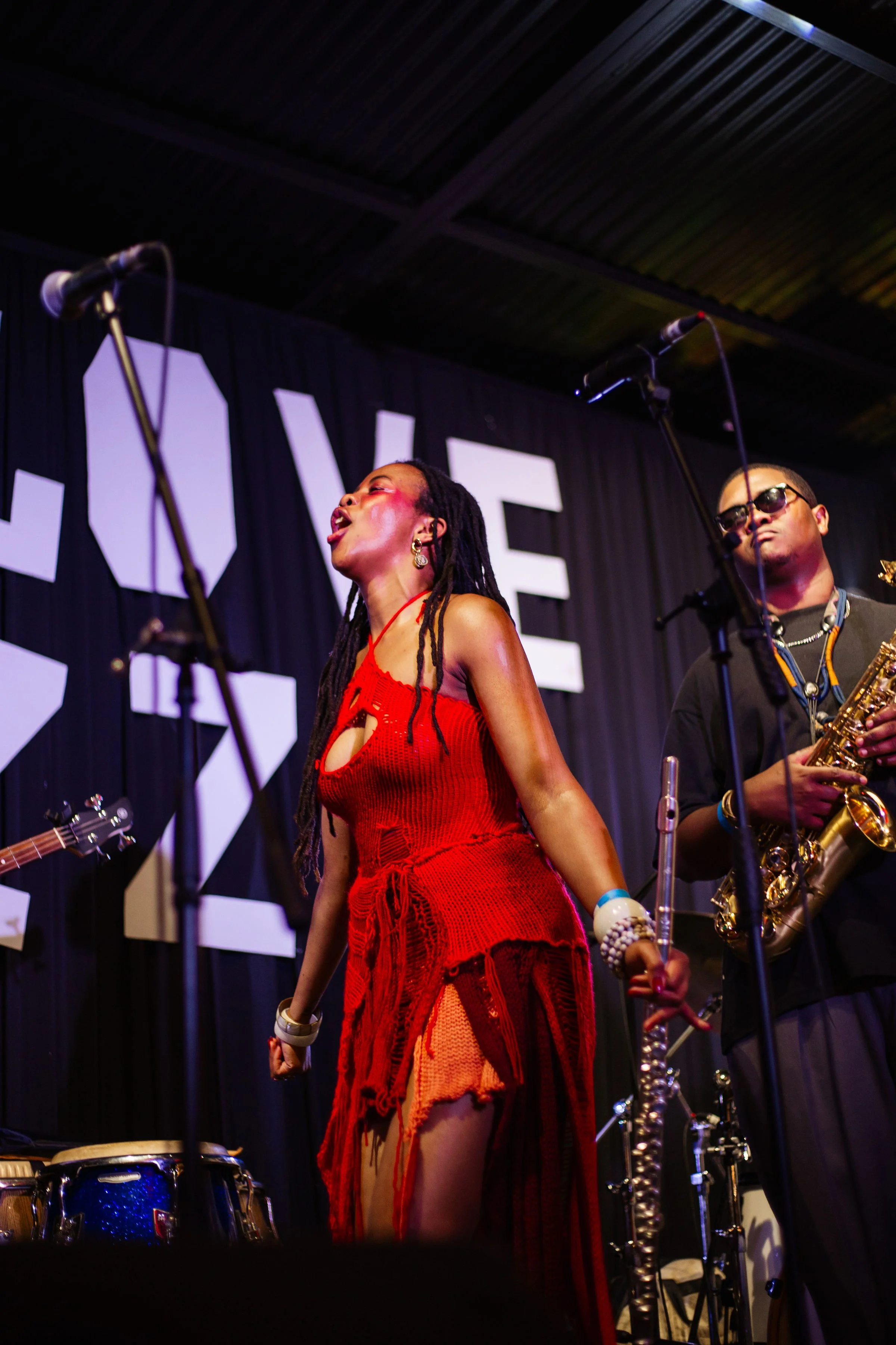 A female singer in a red dress performing on stage with a band, with a male saxophonist beside her, microphones, drums, and musical equipment visible.