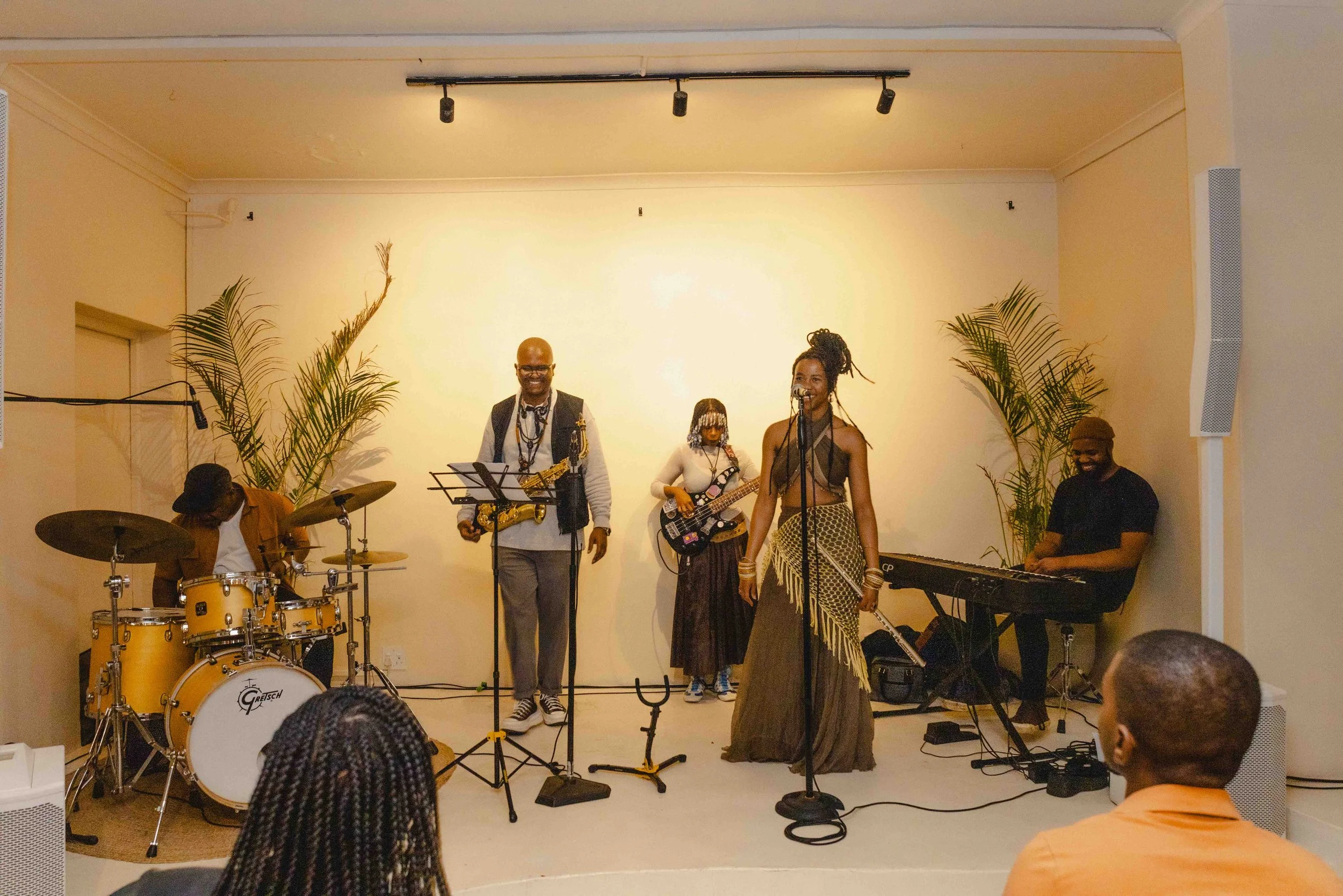 A live music performance featuring a band with several members, including a drummer, a saxophonist, a bass player, a vocalist, and a keyboardist, on a small stage with tropical plants on either side.