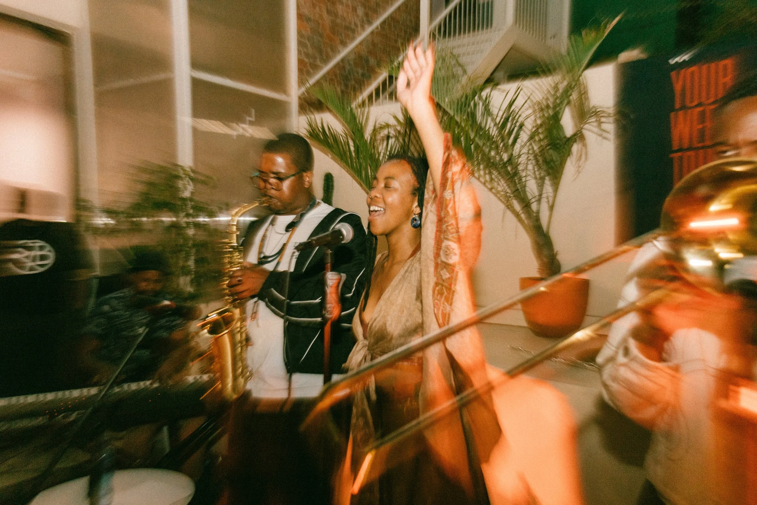 Two women are singing and playing saxophone and trombone at an indoor event, with a woman raising her arm in celebration.