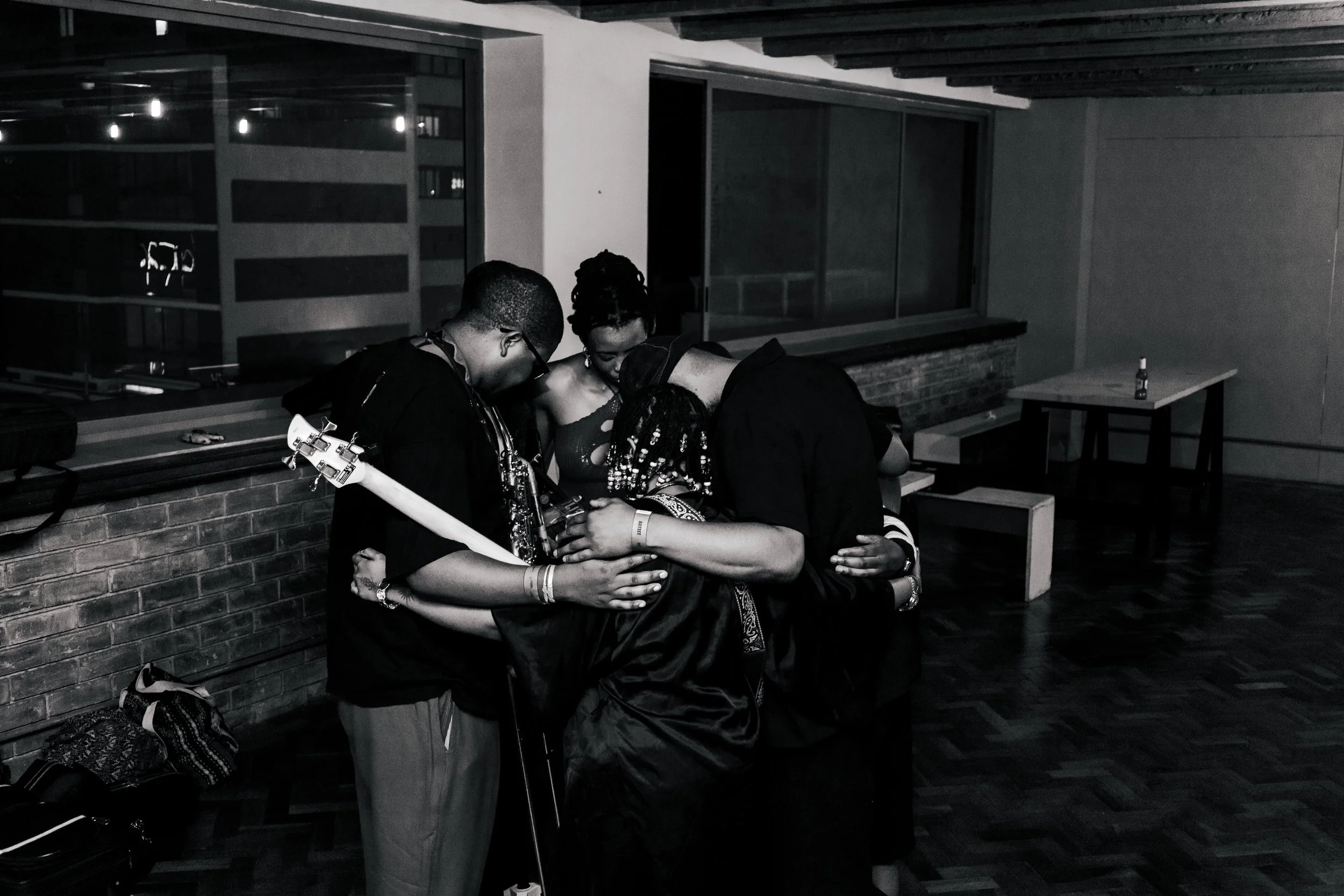 A group of three young women hugging in a dimly lit room with large windows in the background. One woman is holding a guitar, and a bottle is on the table behind them.