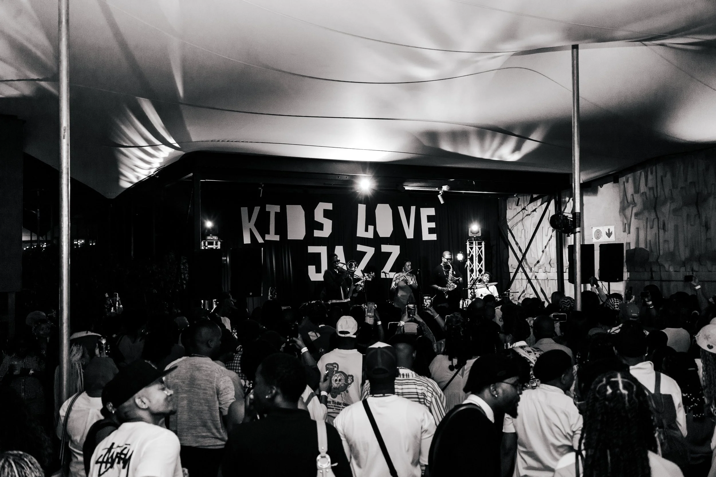 A live musical performance at a venue with an audience. Musicians play instruments on a stage with a banner that says "KIDS LOVE JAZZ." The scene is in black and white.