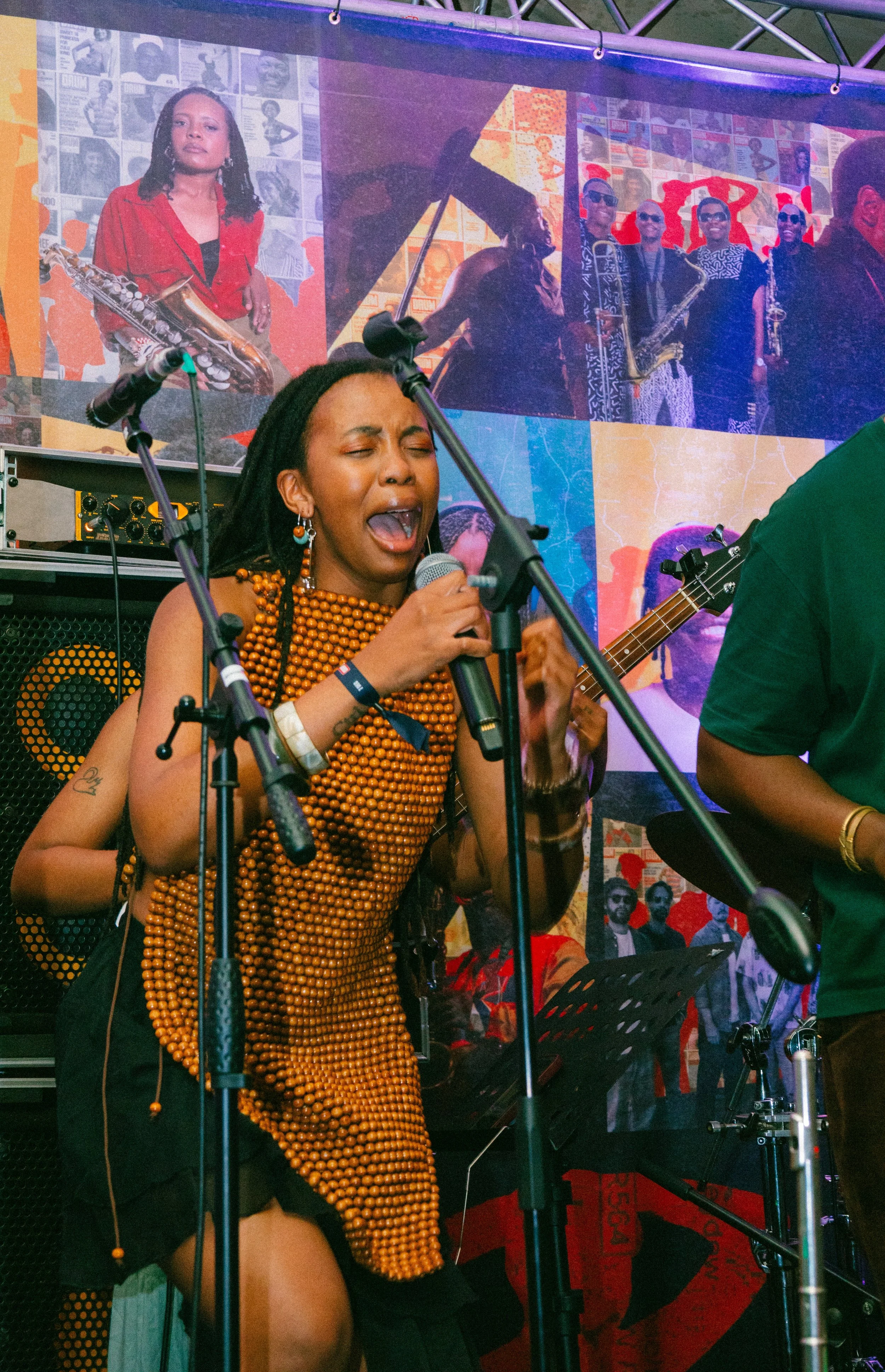 A woman singing passionately into a microphone during a live musical performance. She is wearing a beaded orange dress and jewelry, with musical equipment and a large colorful poster in the background depicting various people and images.