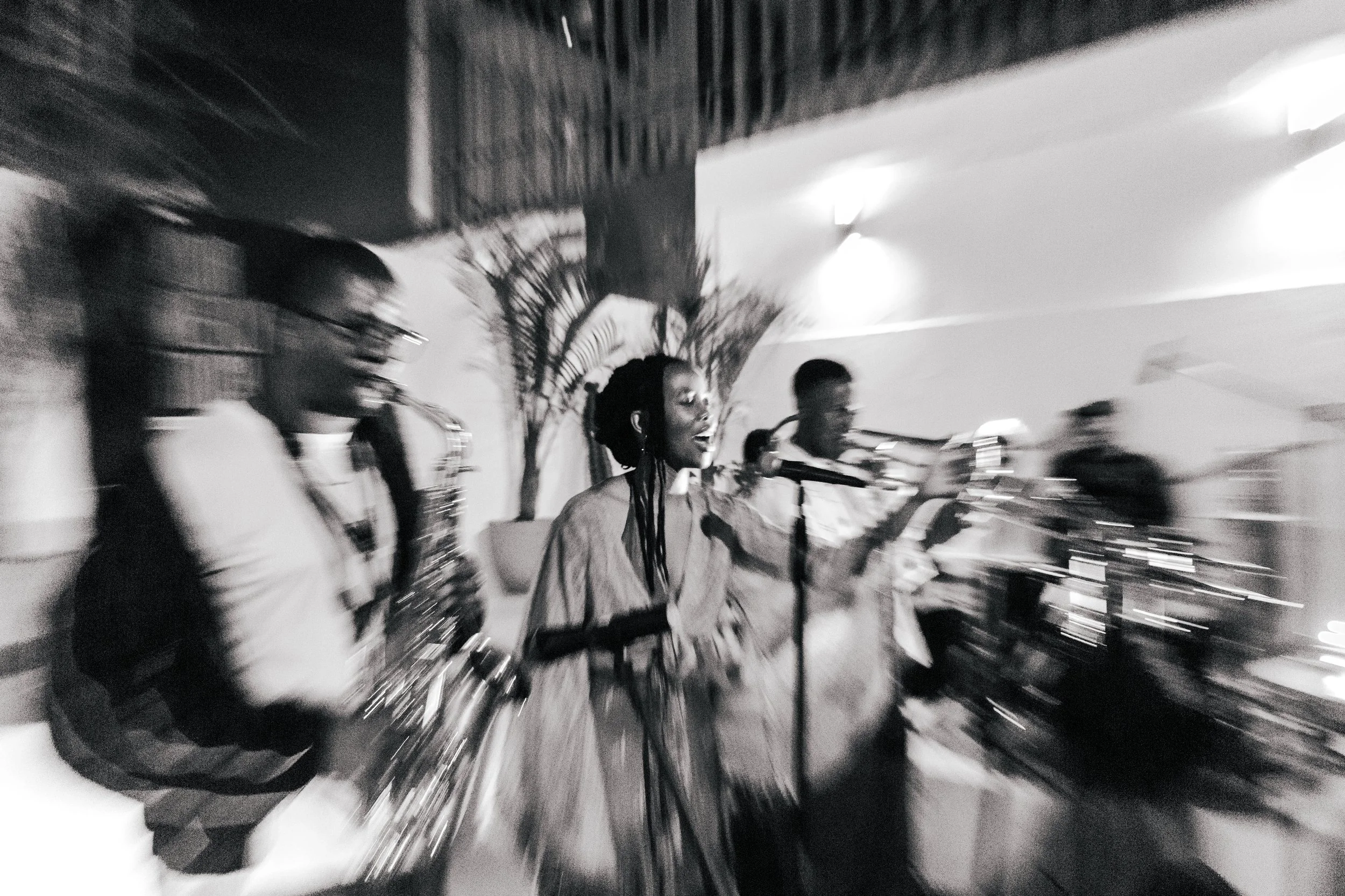 Black and white photo of three women singing and playing musical instruments, with one woman in the center singing into a microphone. The scene appears to be indoors with lighting and a minimal background.