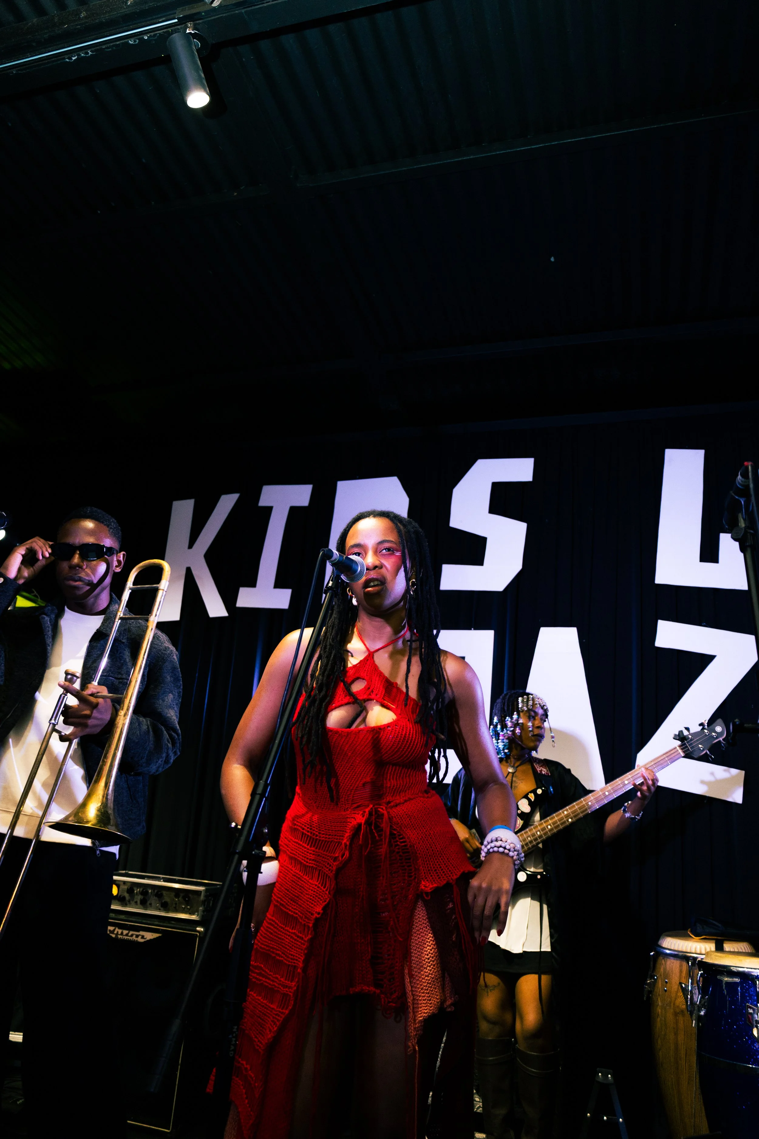 A live music performance with several artists playing instruments and singing on stage, including a woman in a red dress singing into a microphone and a man with sunglasses playing a trombone. The stage has a black curtain backdrop with large white l