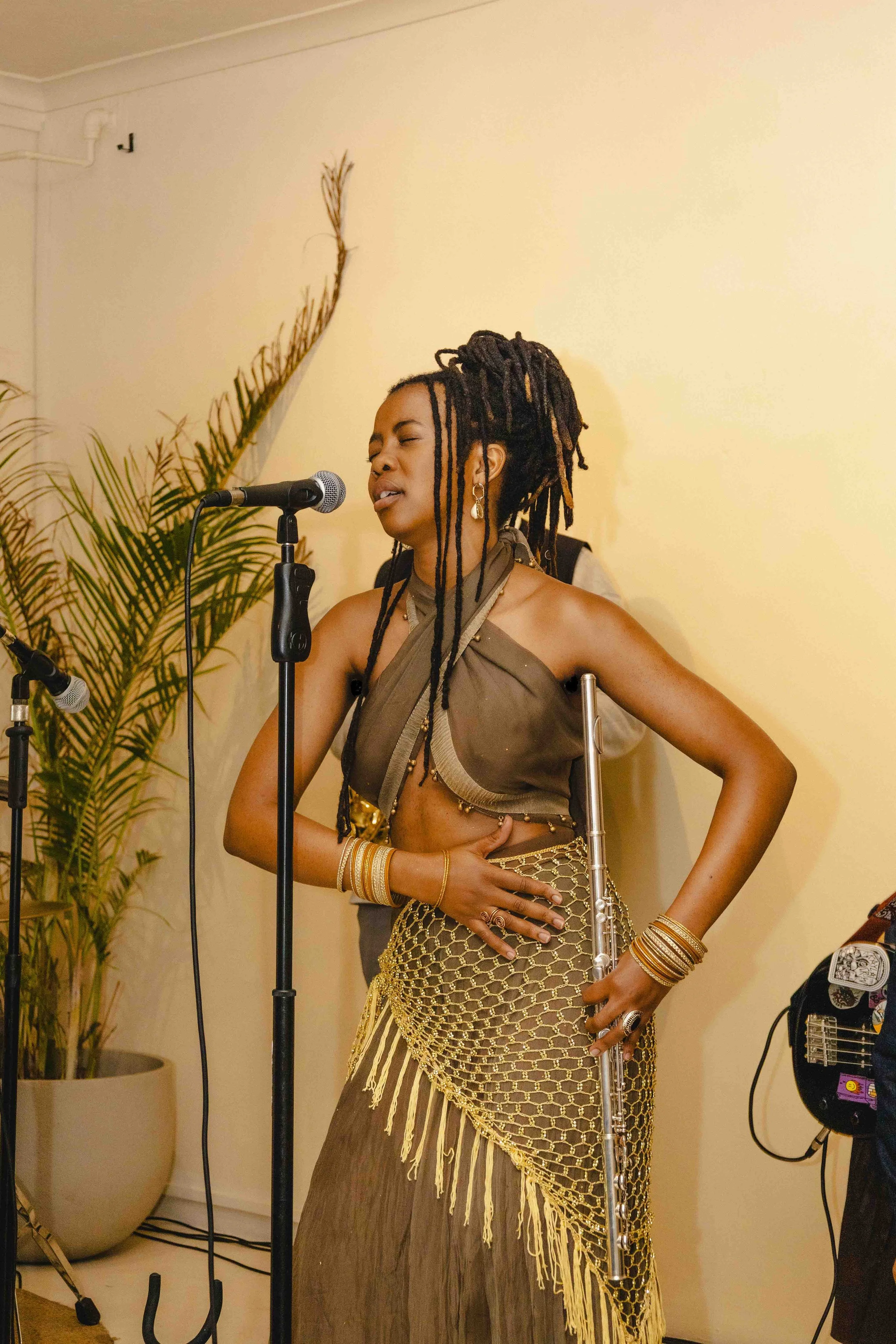 A woman with long dreadlocks singing into a microphone, dressed in a brown and gold belly dance costume with jewelry, holding a flute, standing in front of a beige wall and potted plants.