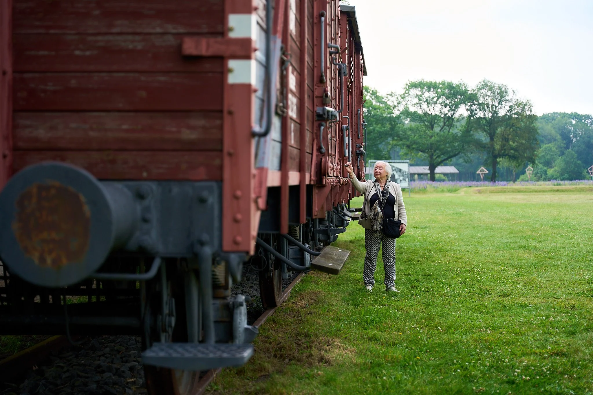 An elderly woman standing outdoors beside a red vintage train, touching the train's side, with green trees and a grassy field in the background.