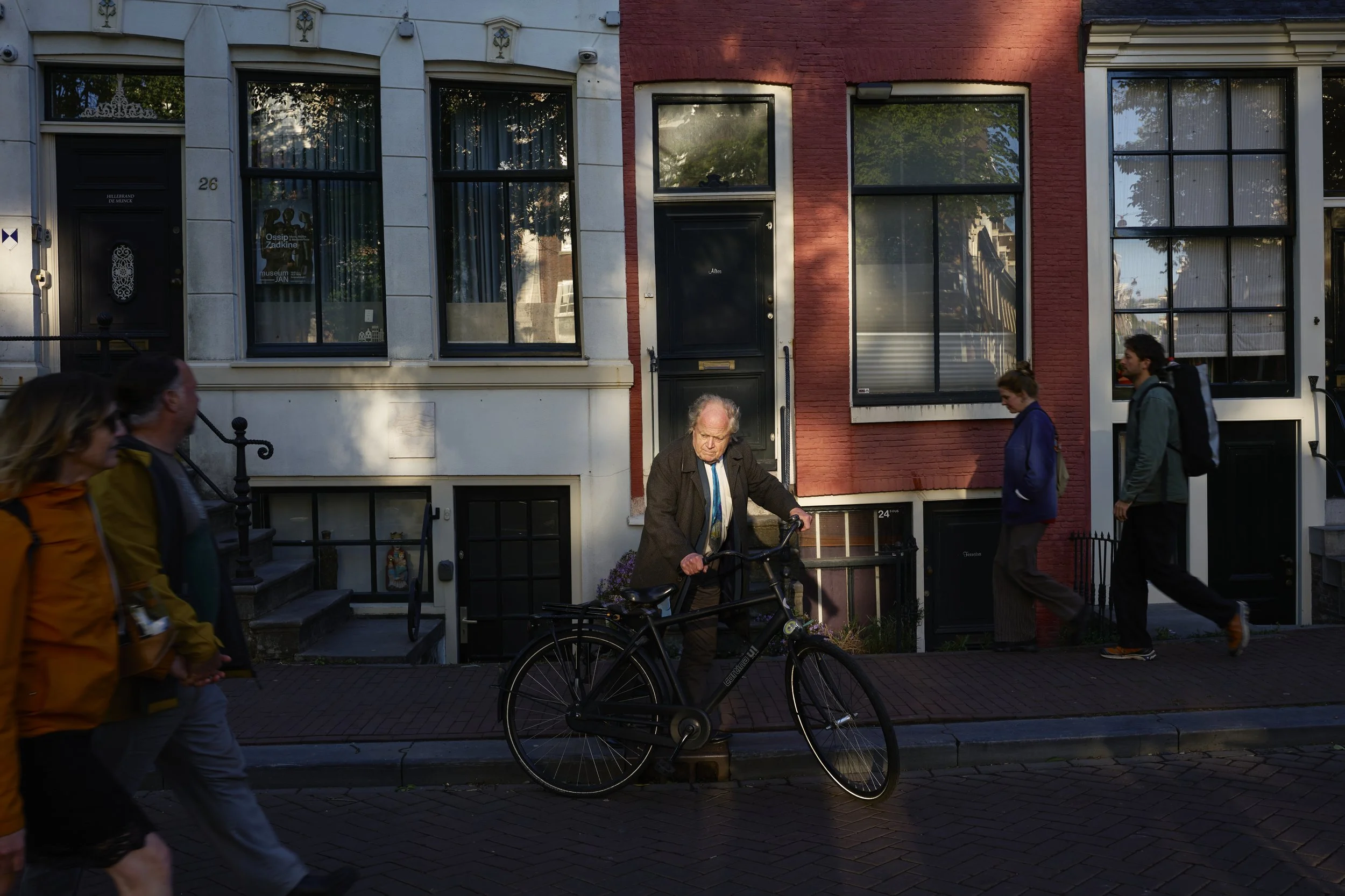 An elderly man with long gray hair and glasses, wearing a dark jacket, stands with his bicycle on a city sidewalk. Several pedestrians walk past in front and behind him. In the background are row houses with large windows, some reflecting trees and s