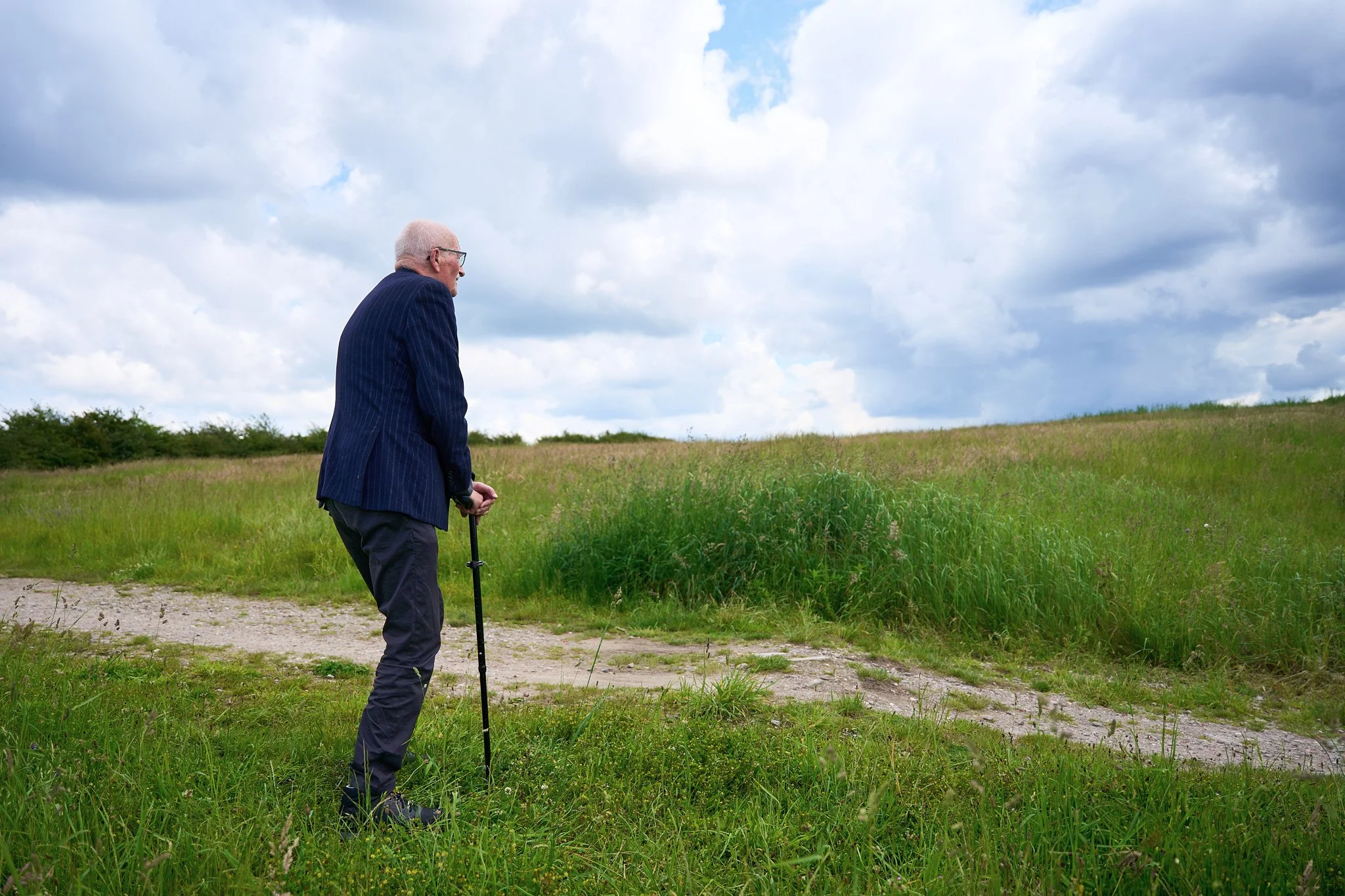 An elderly man in a dark pinstripe suit walks on a grassy trail with a cane in a vast, open field under a cloudy sky.
