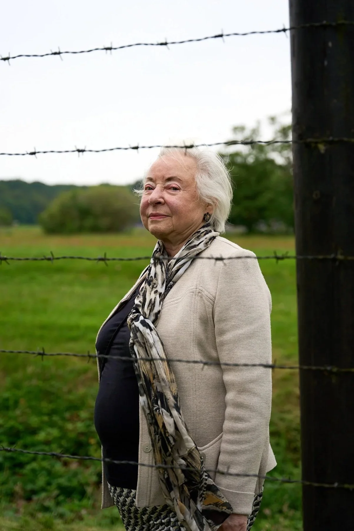 An elderly woman with white hair standing outdoors near a barbed wire fence, with a green field and trees in the background.